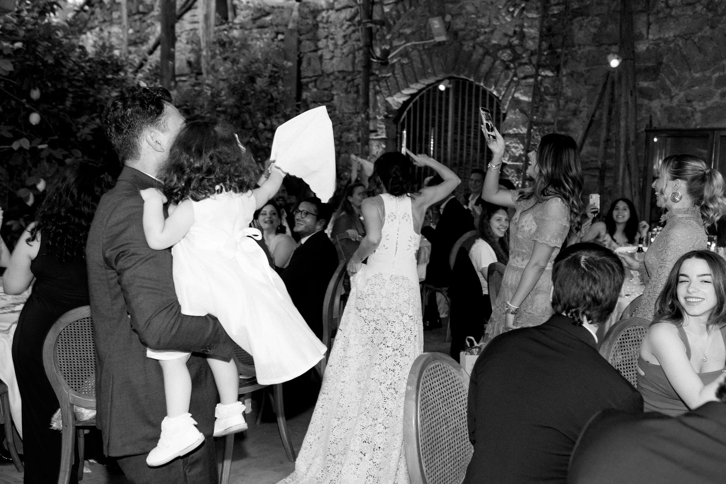 Una scena di festa con persone sedute e in piedi, alcune che scattano foto, e una ragazza con un vestito lungo in piedi al centro, in un ambiente rustico con pareti di pietra.