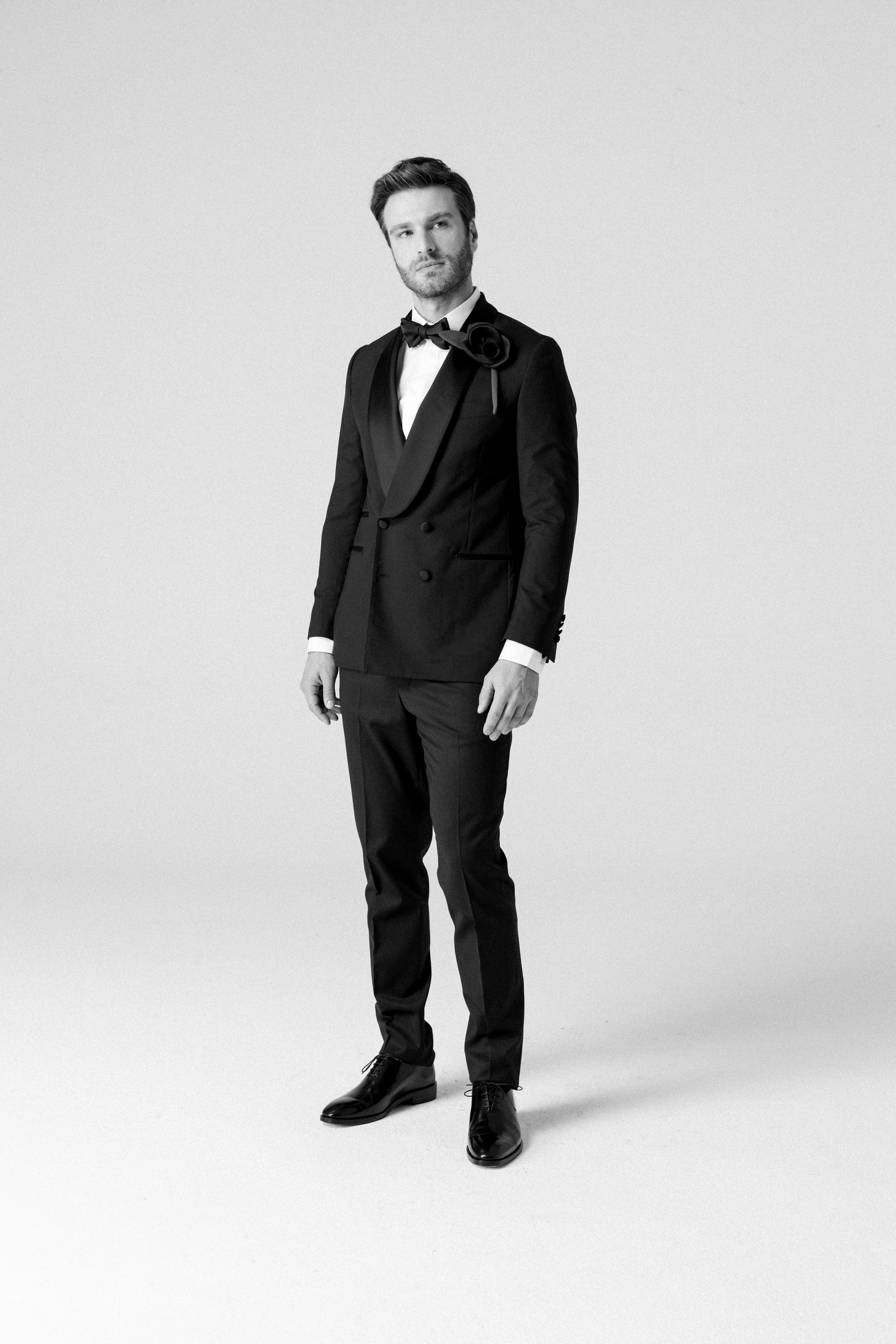 Uomo in abito da sera nero con cravatta e fiocco, posa su sfondo bianco.