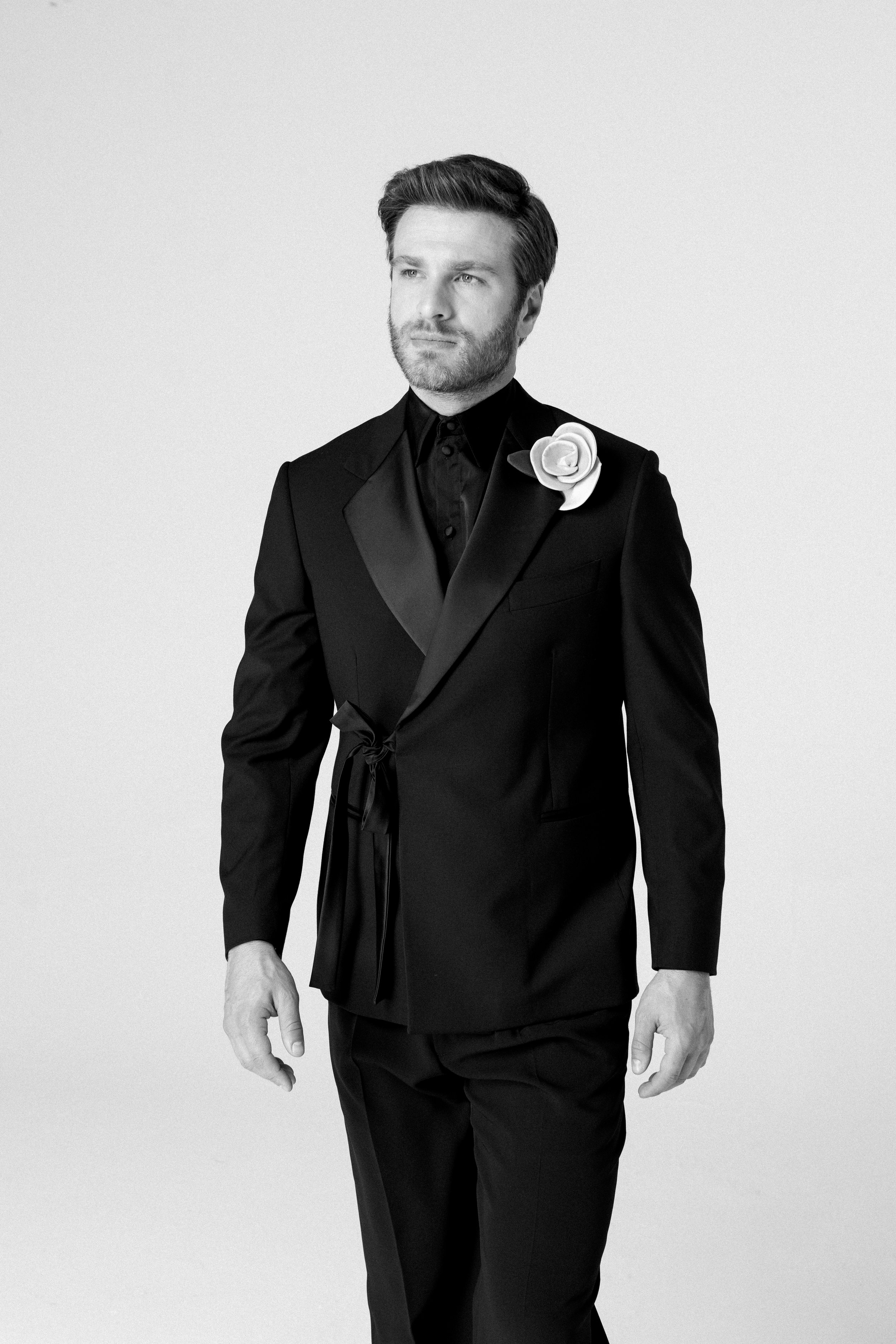 Uomo in abito nero con fiore bianco sulla spalla, in posa frontale, in bianco e nero.