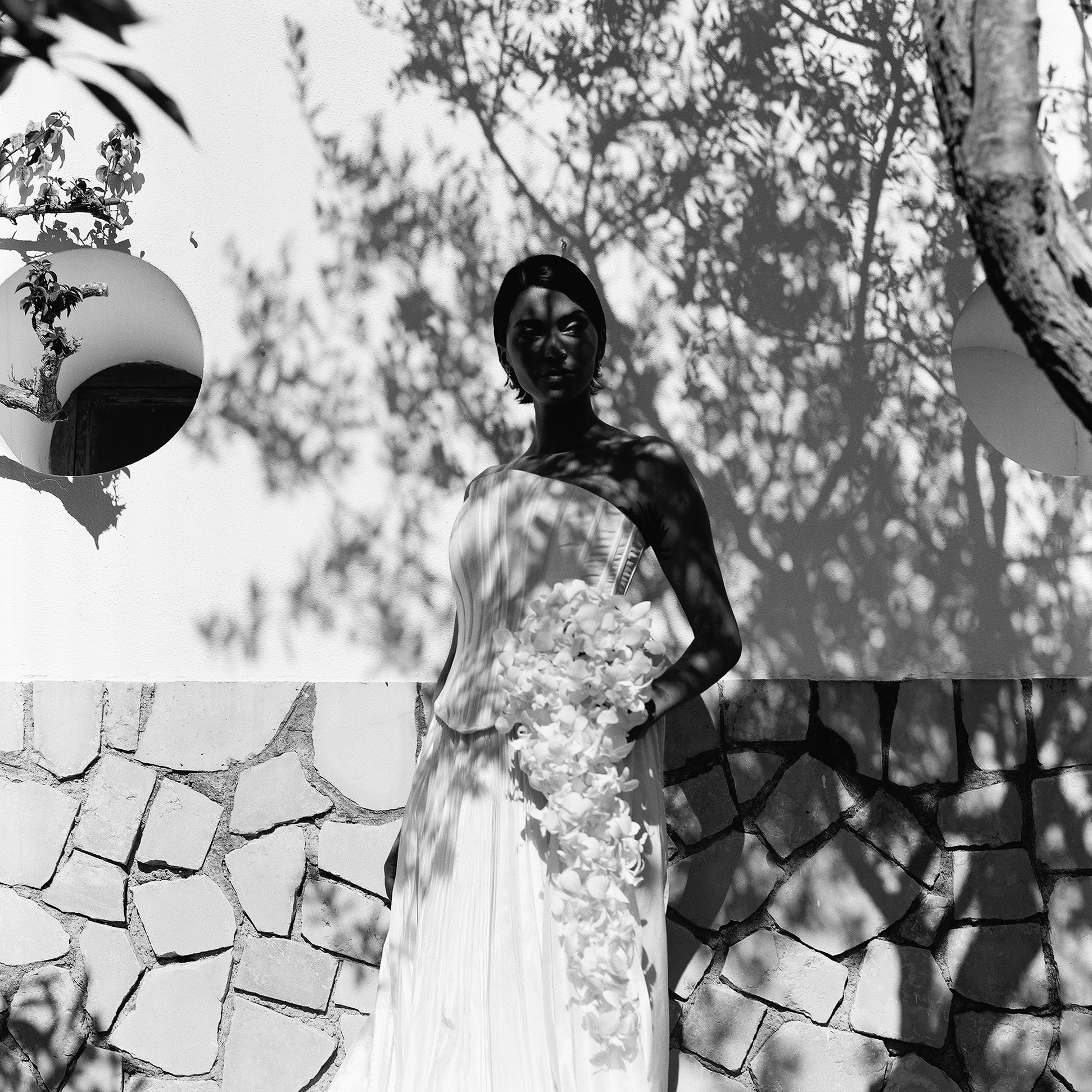 Una donna indossa un abito lungo bianco e tiene un mazzo di fiori, con ombre di foglie e rami che proiettano ombre sul suo volto e sul corpo, in un ambiente esterno con parete di pietra e decorazioni sferiche.