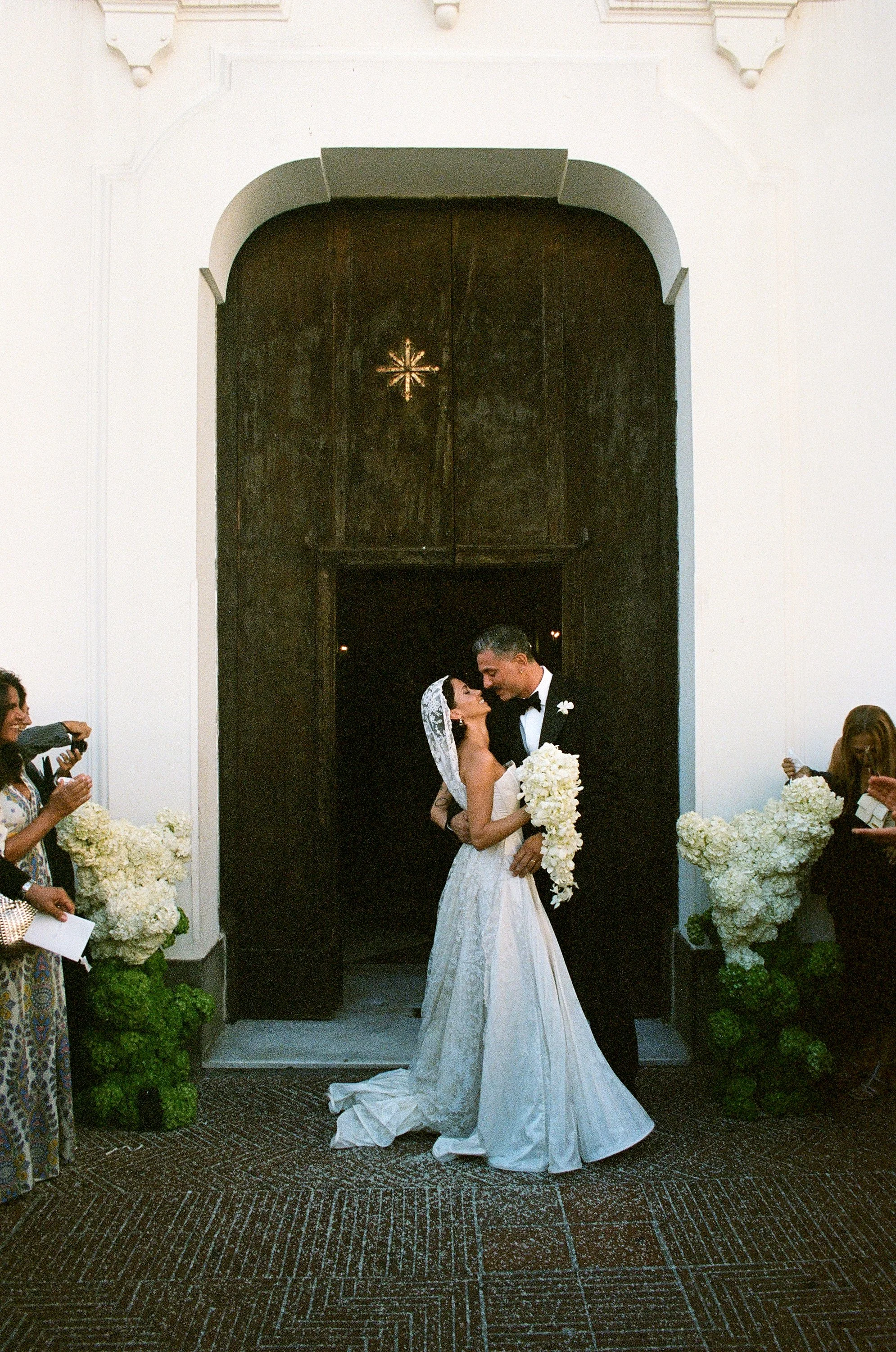 Una coppia di sposi si bacia all'uscita di una chiesa, circondati da amici e decorazioni floreali bianche.