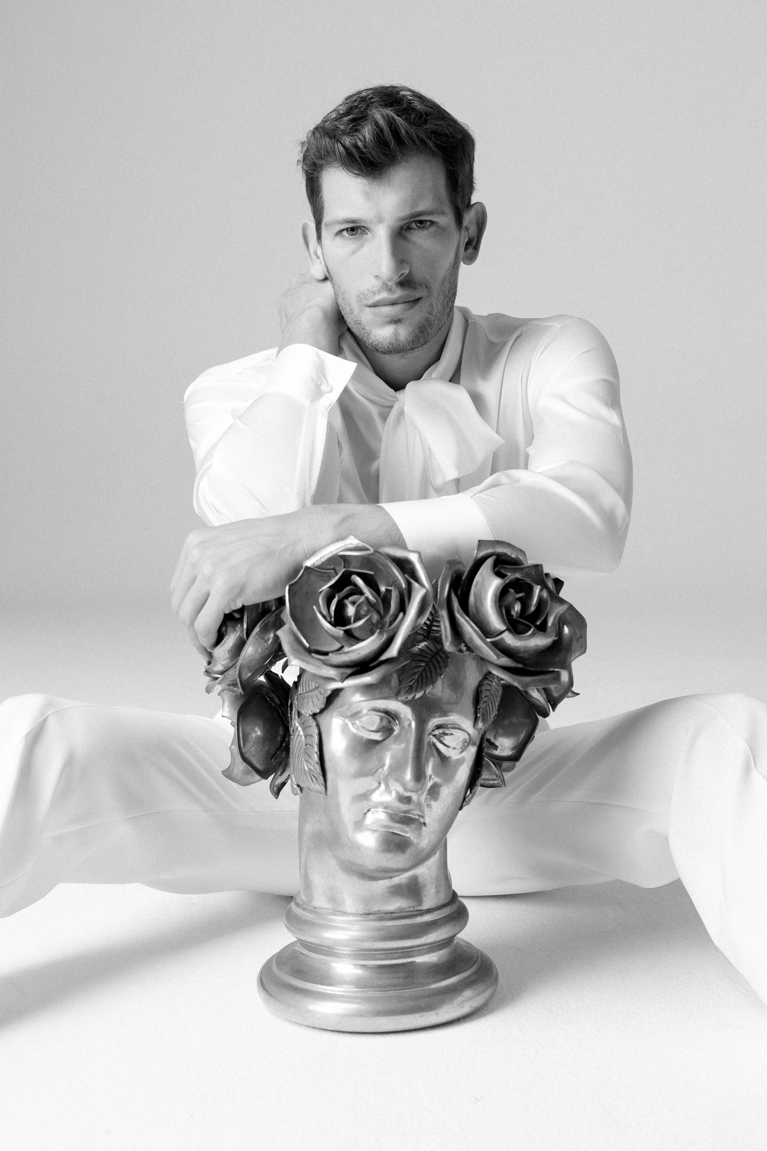 Uomo seduto di fronte a una testa scolpita di un volto umano con fiori di rose come acconciatura, in uno stile artistico e moderno, in bianco e nero.