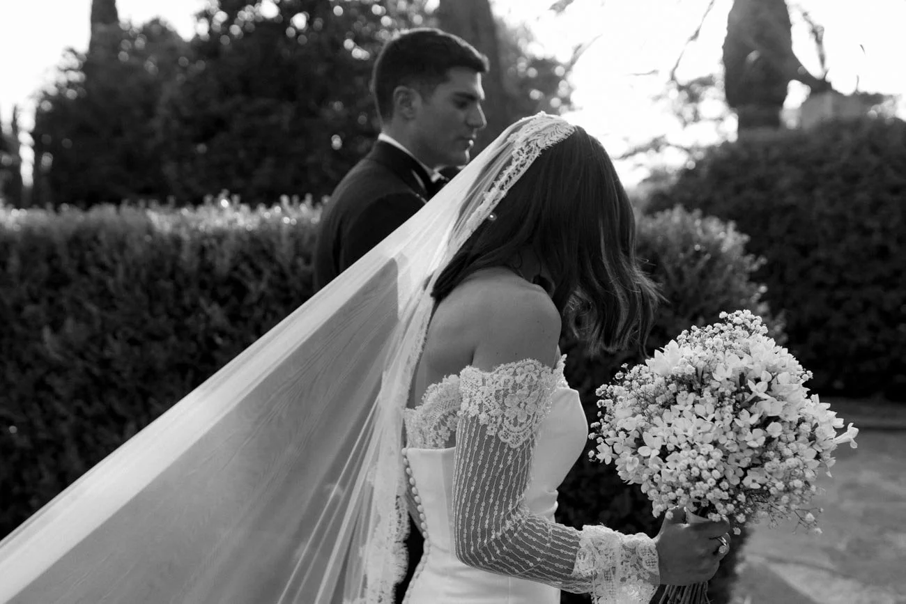 Una coppia di sposi in abiti da matrimonio, la donna tiene un bouquet di fiori mentre lui si trova sullo sfondo, in un giardino.