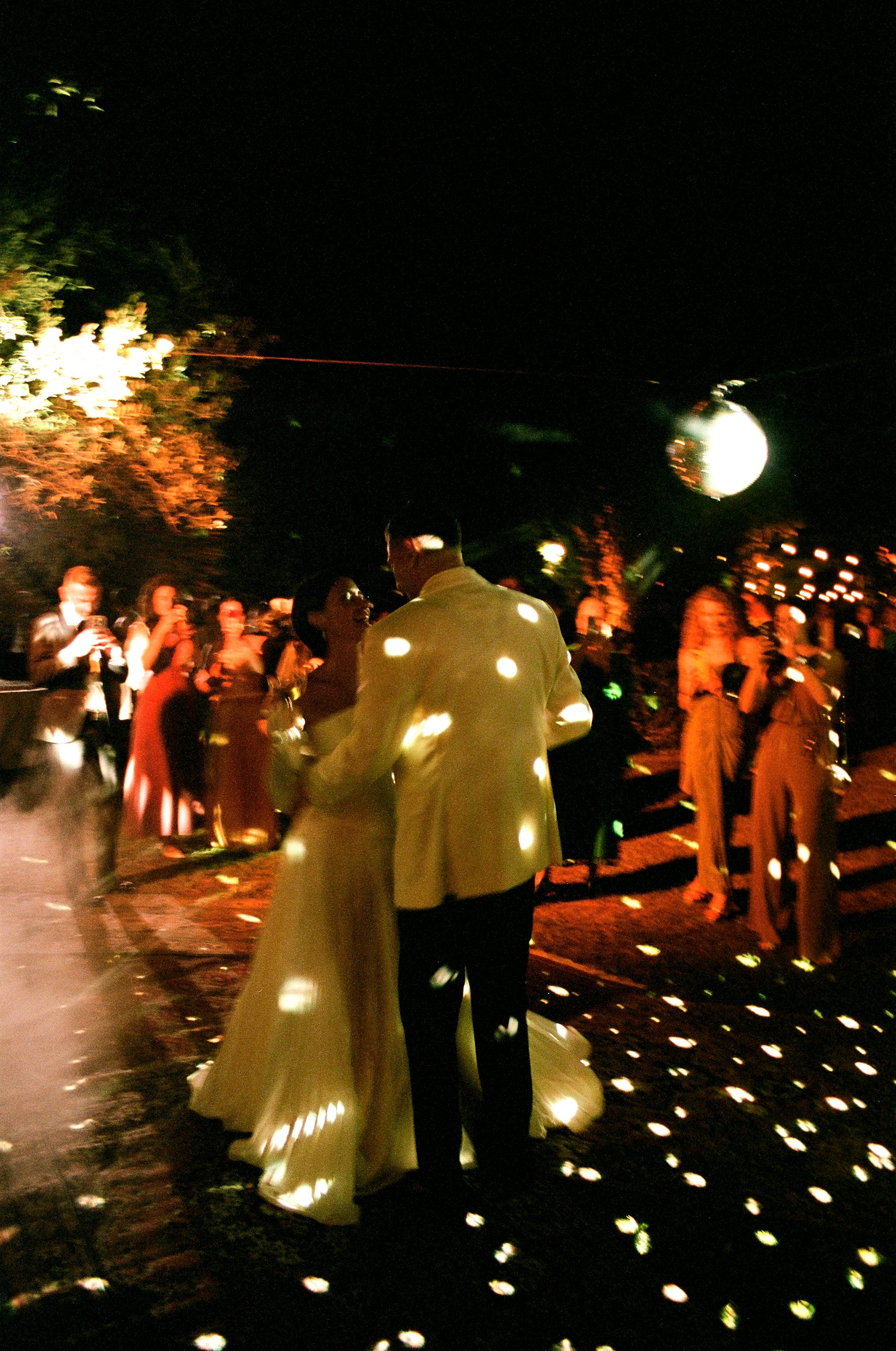 Una coppia di sposi che ballano alla sera durante un matrimonio all'aperto, circondati da amici e ospiti, con luci soffuse e una palla da discoteca che riflette la luce.