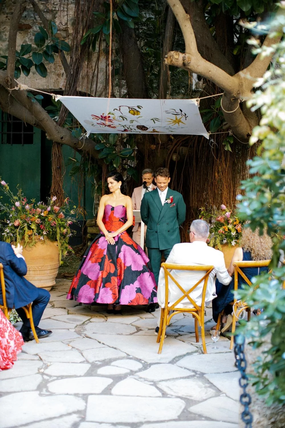 Cerimonia di matrimonio all'aperto con coppia di sposi sotto un grande albero, persone sedute, fiori e decorazioni colorate.