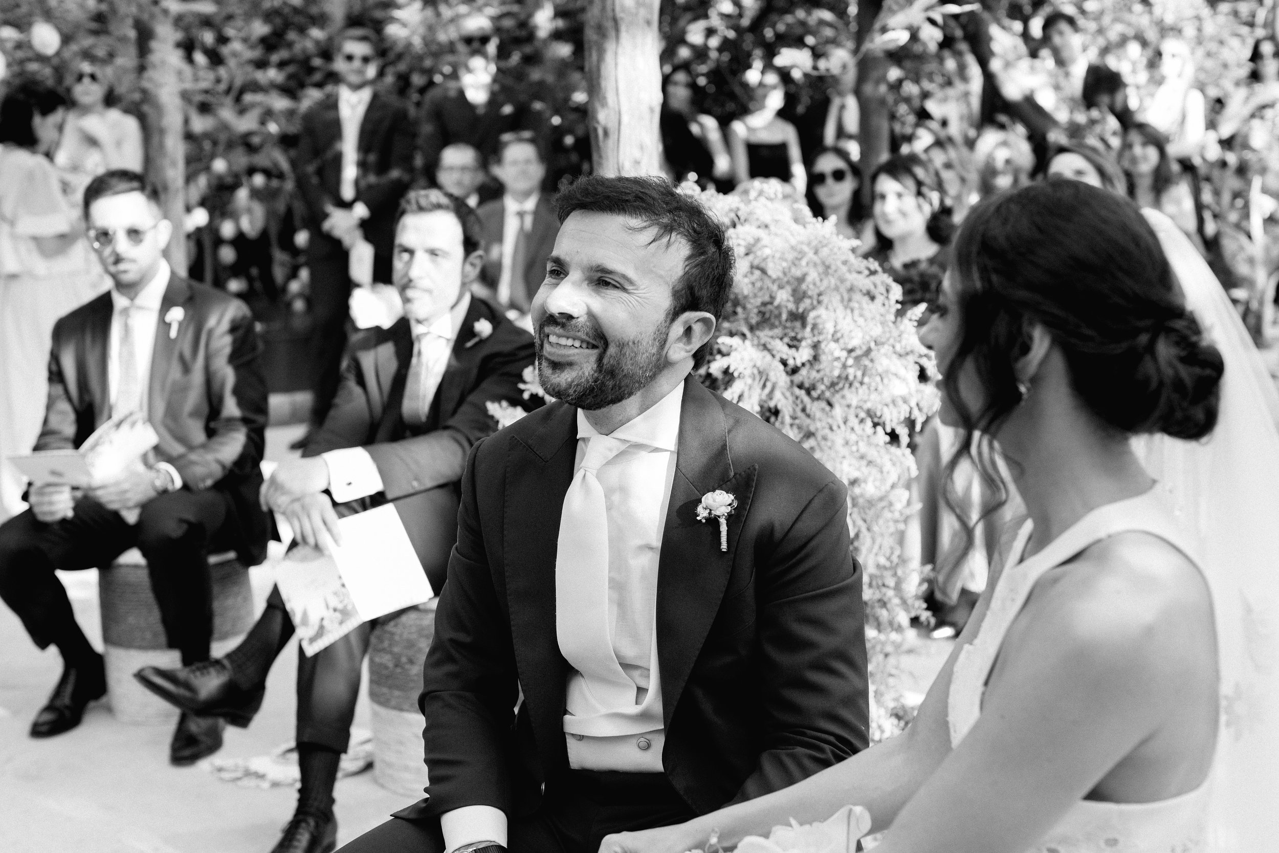 Una coppia di sposi in coincidenza di un matrimonio, seduti insieme, con invitati in background, in un'ambientazione all'aperto, in bianco e nero.