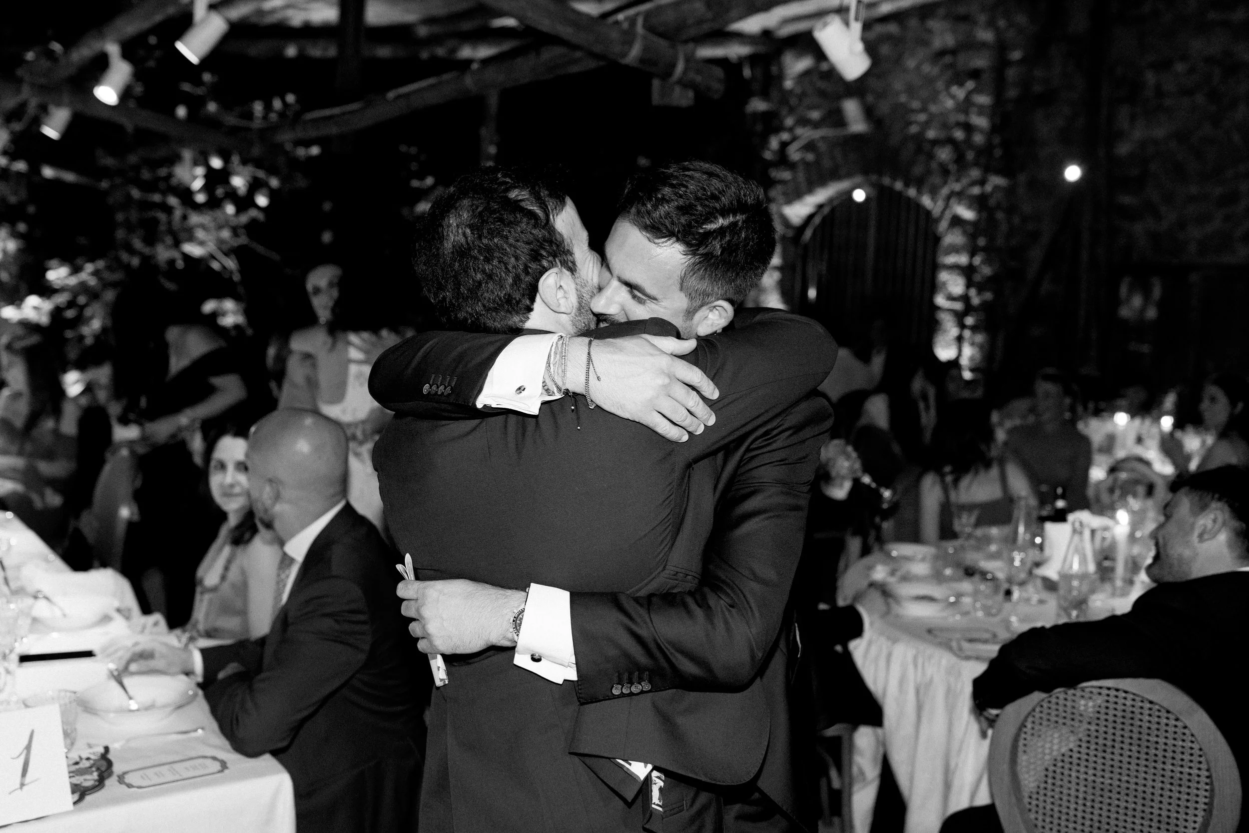 Due uomini in abiti eleganti si abbracciano tendendosi per scambiarsi un bacio in un evento sociale, con molte persone sedute a tavoli in background.