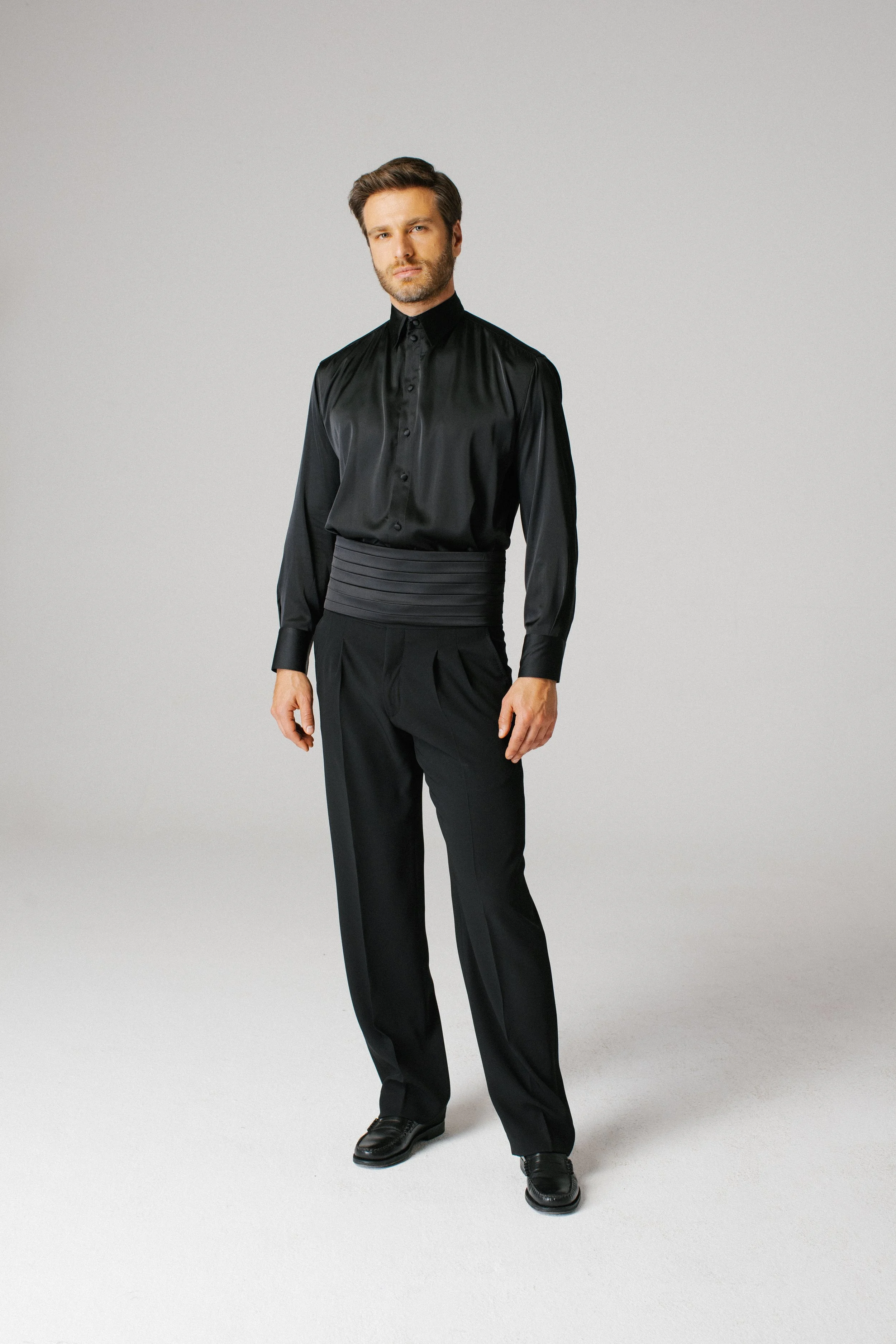 Uomo vestito con abiti eleganti neri, camicia di seta, pantaloni e scarpe eleganti in piedi contro uno sfondo grigio chiaro.