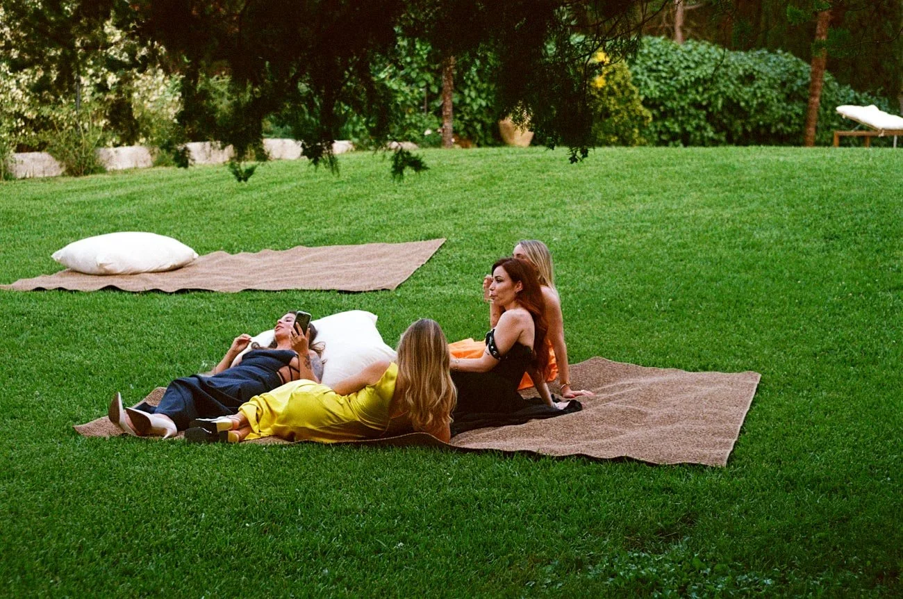 Gruppo di donne che si rilassa su coperte di paglia stese sull'erba in giardino, alcune sdraiate e altre sedute, alcune che conversano e altre che guardano il telefono, con spazio verde e alberi sullo sfondo.