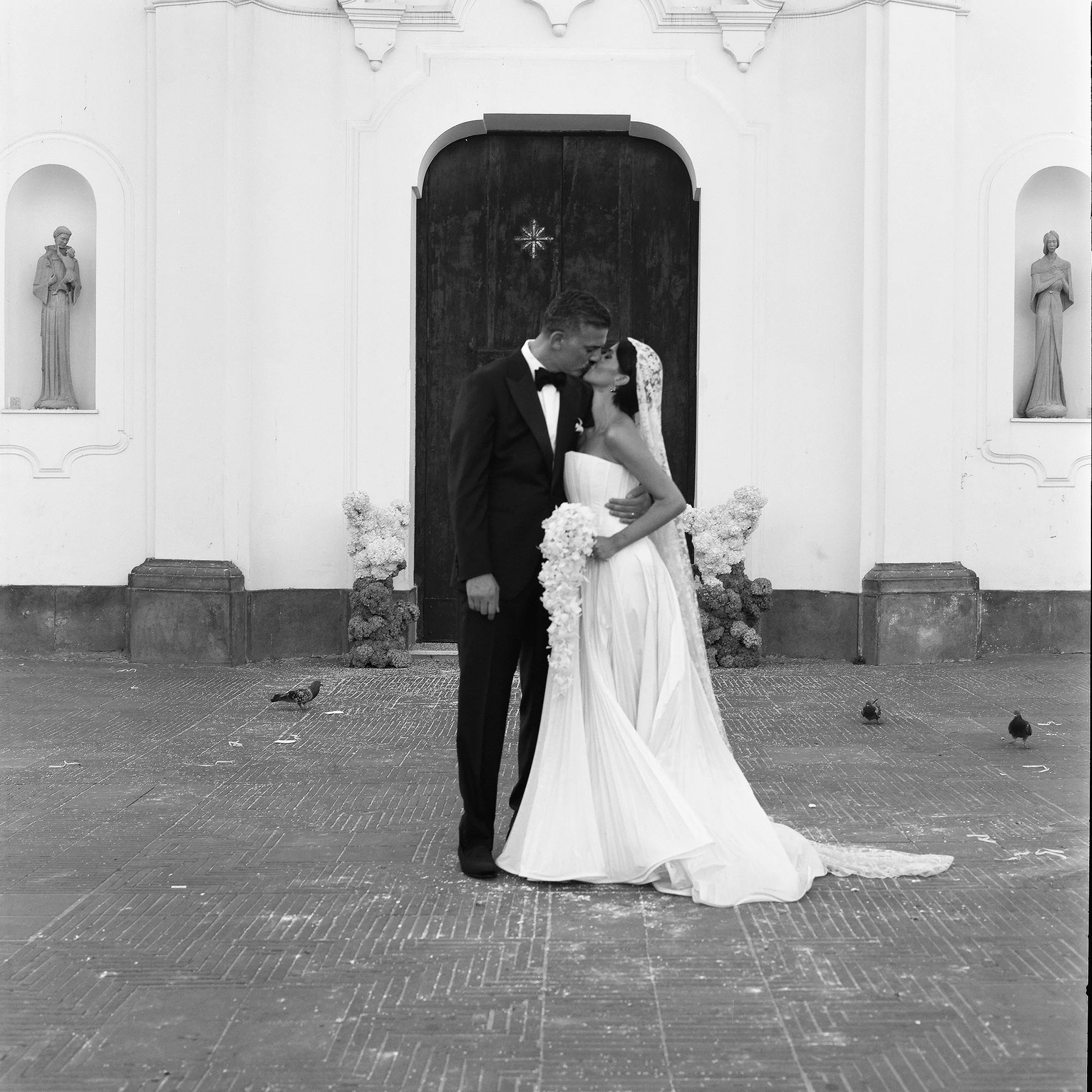 Una coppia di sposi si bacia davanti a una porta di chiesa, con decorazioni floreali ai lati, in bianco e nero.