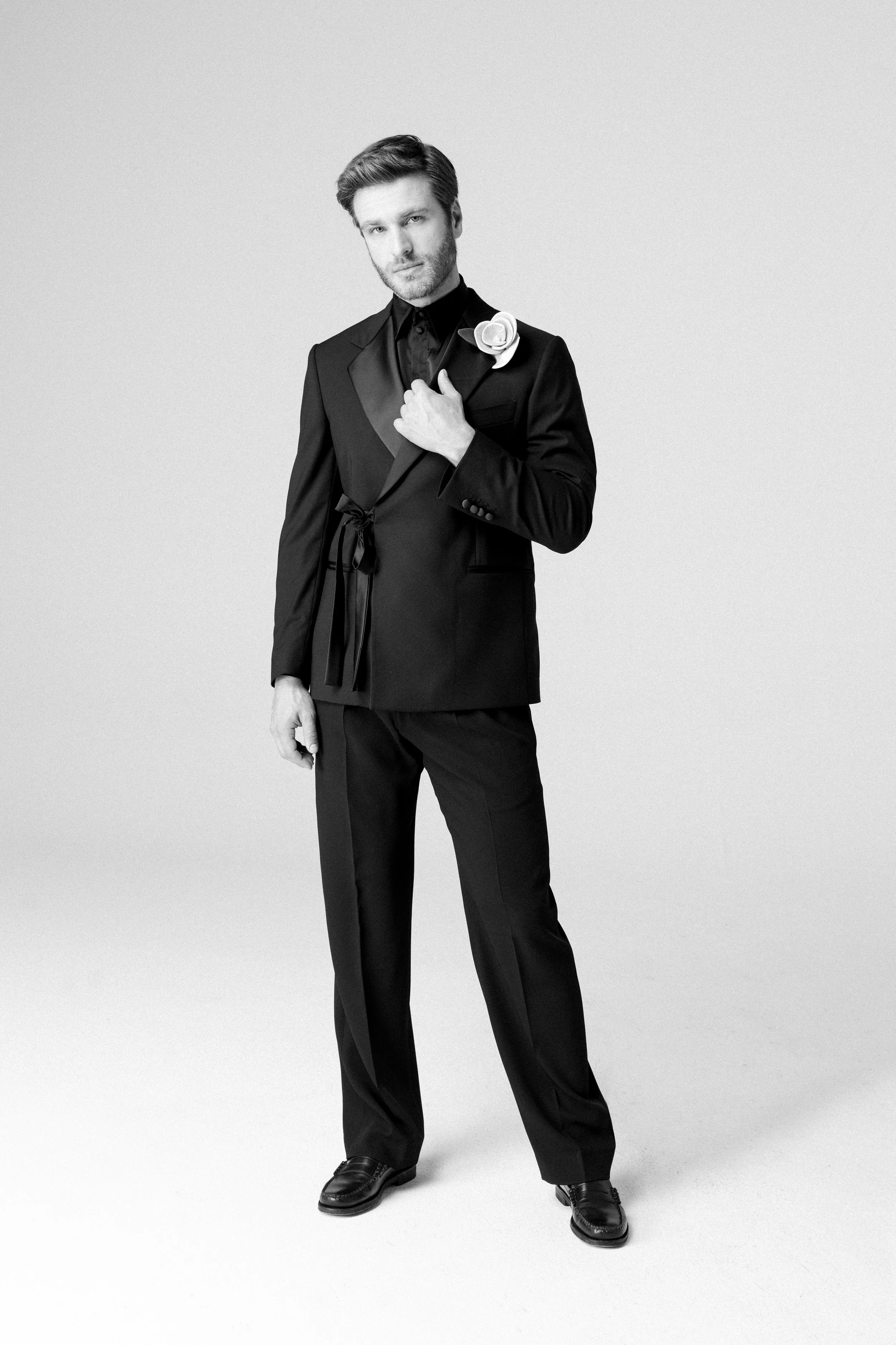 Uomo in abbigliamento elegante nero, posa con una mano sul petto, sfondo neutro.