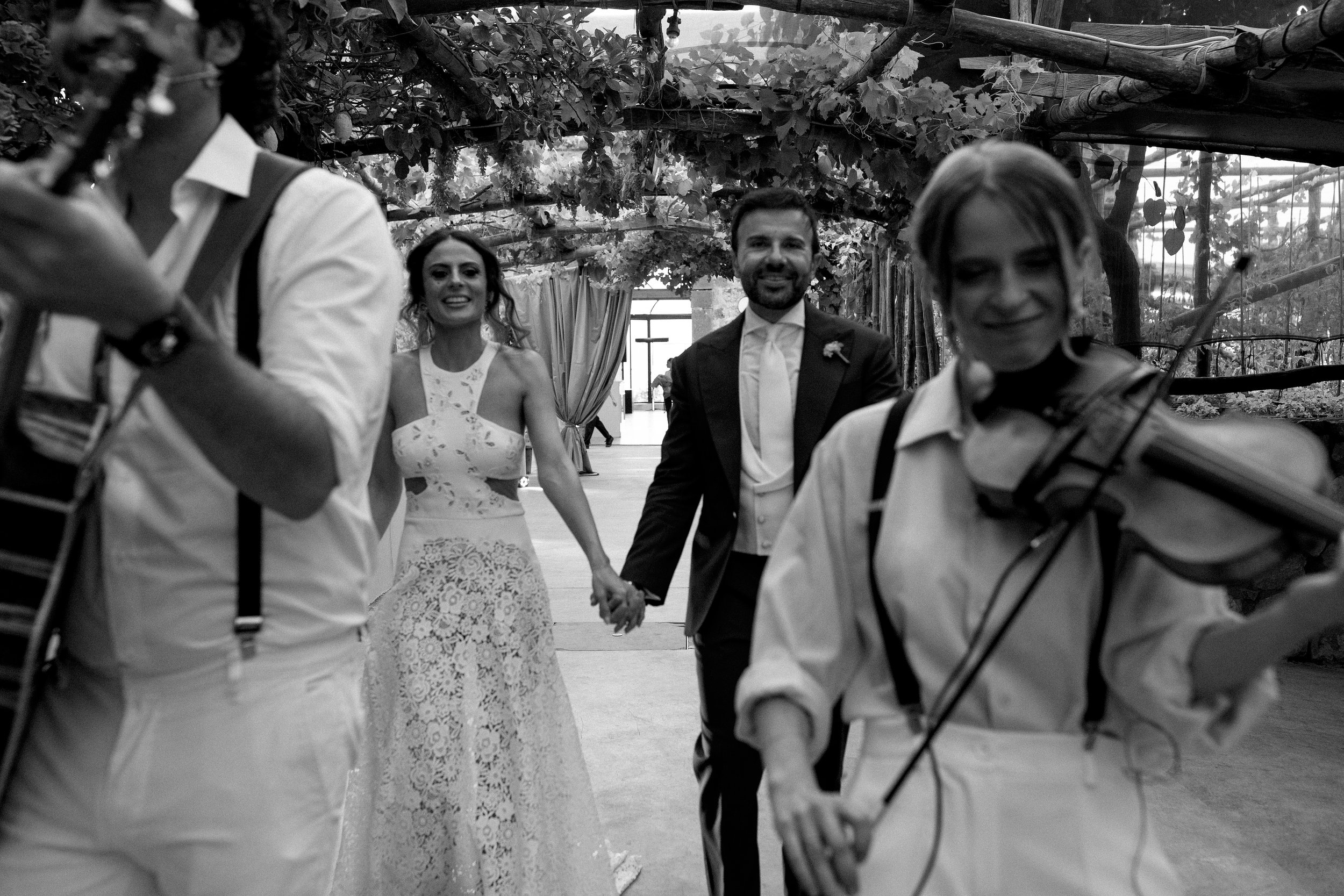 Una coppia di sposi tiene le mani di fronte a loro, camminando tra musicisti che suonano, in un ambiente naturale.