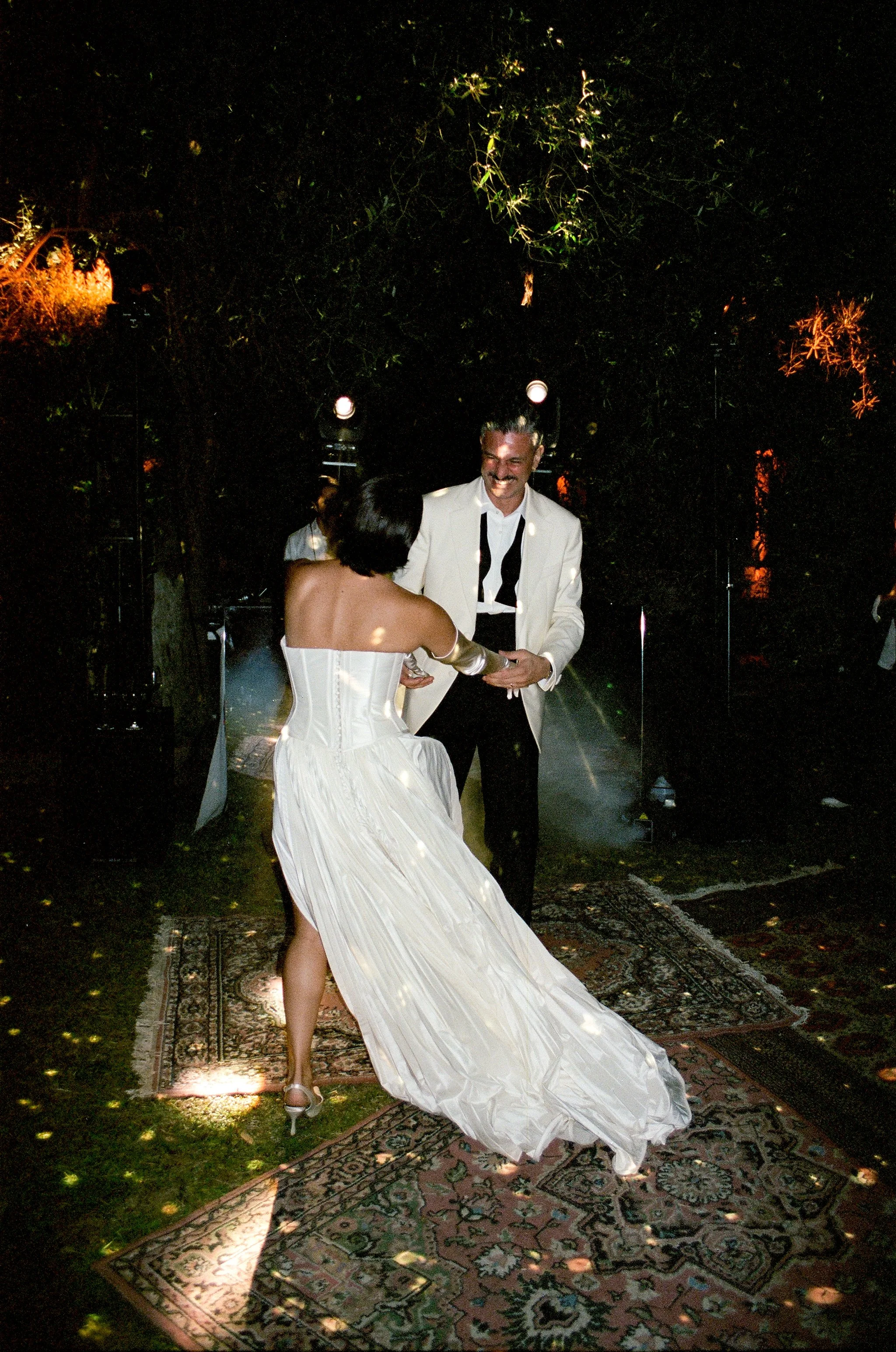 Una coppia che balla durante una festa in un ambiente all'aperto, con luci soffuse e tappeti sul prato.