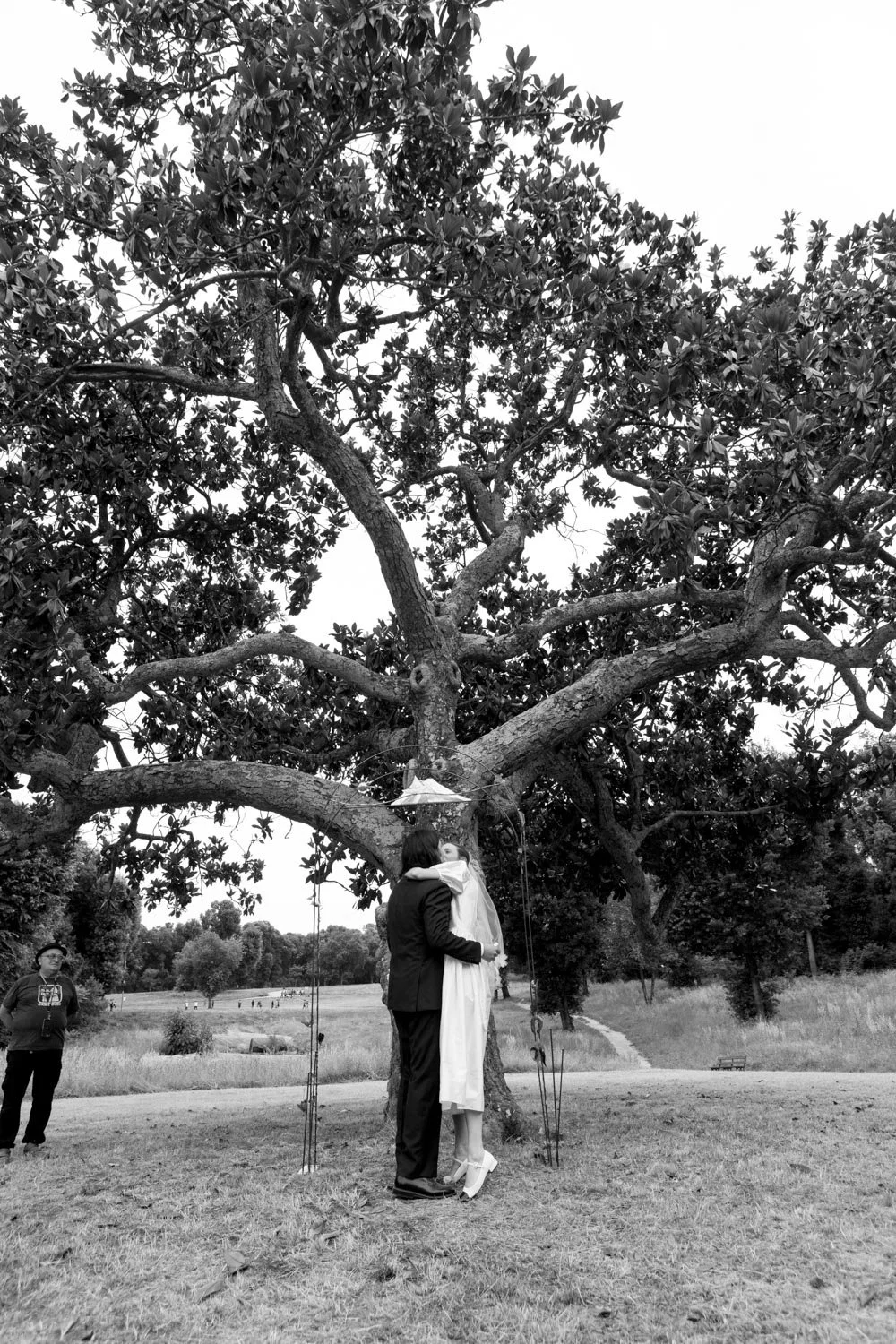 Una coppia di sposi si bacia sotto un grande albero in un parco. Una persona a sinistra osserva. L'immagine è in bianco e nero.