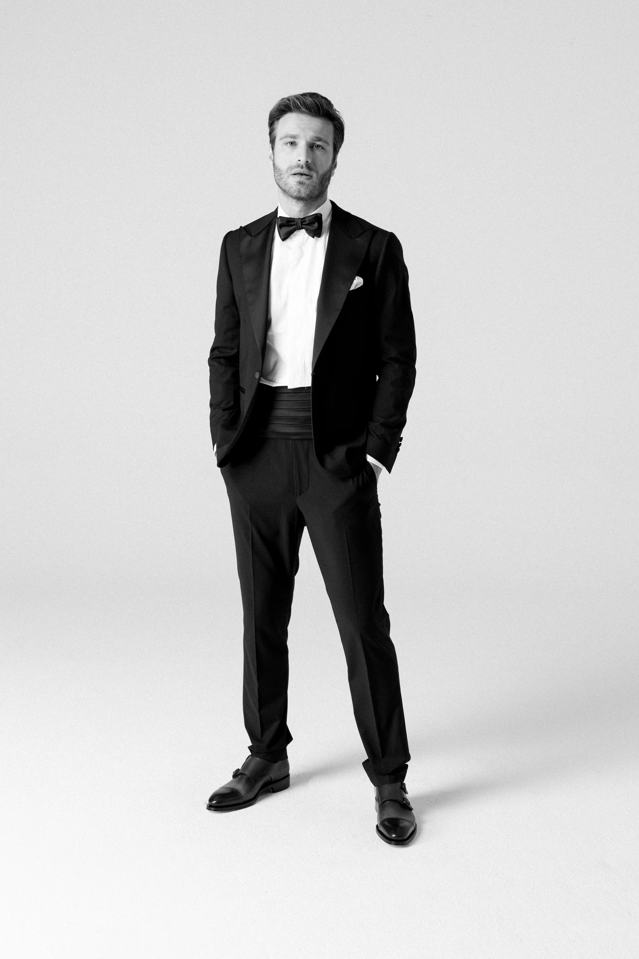 Uomo vestito con smoking nero e papillon nero, posa con le mani in tasca, sfondo bianco.
