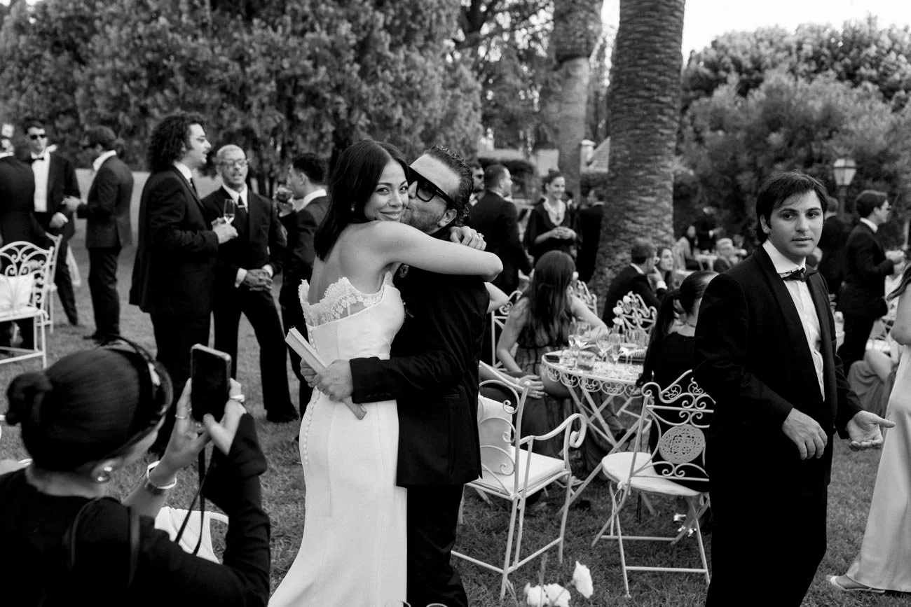 Una coppia si abbraccia sorridente durante una cerimonia di matrimonio all'aperto, con altri invitati in sfondo. È un'immagine in bianco e nero.