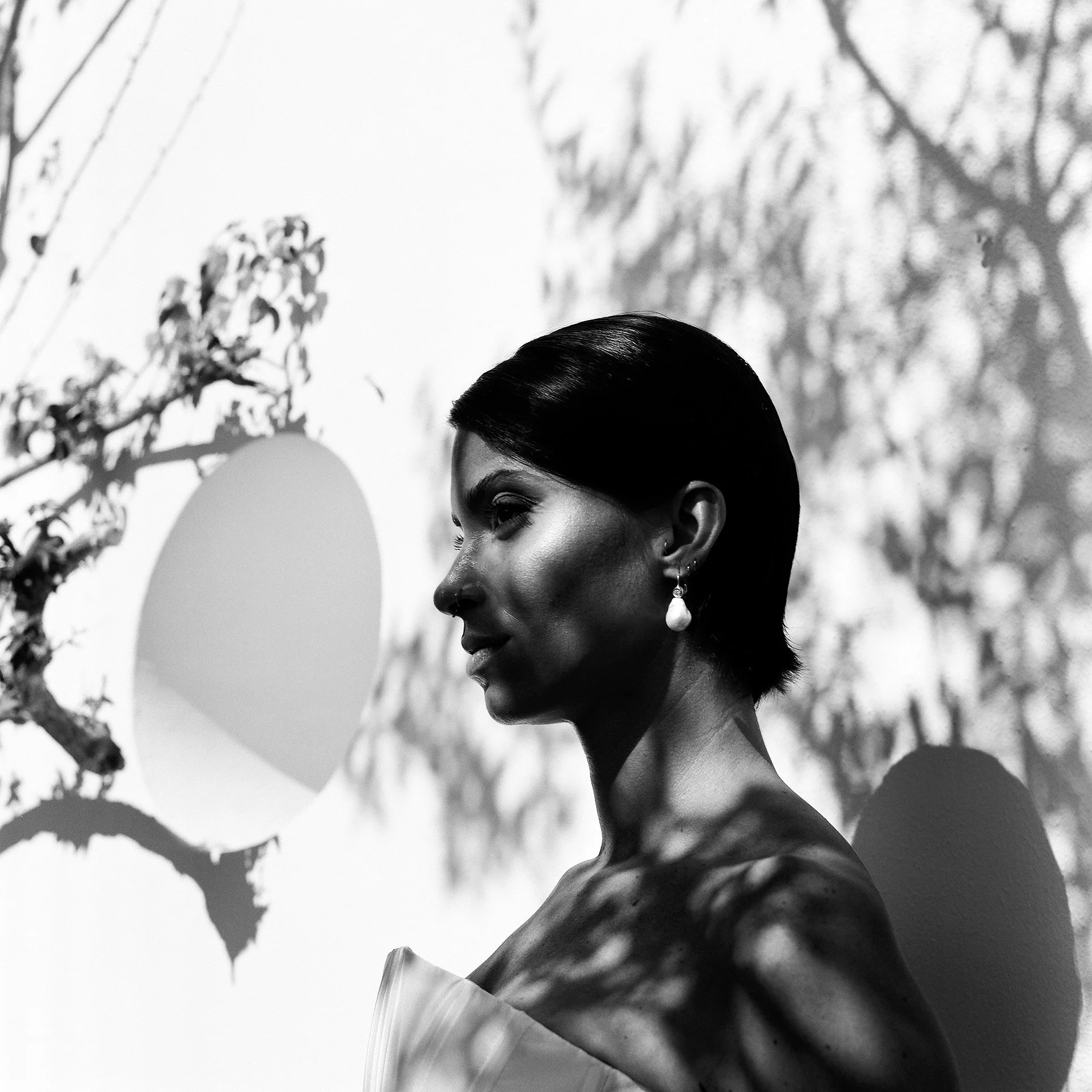 Ritratto in bianco e nero di una donna con capelli neri corti, che indossa un abito senza spalline e orecchini pendenti a goccia, con ombre di rami e foglie sullo sfondo che creano un gioco di luci e ombre sulla sua pelle e sulla parete.