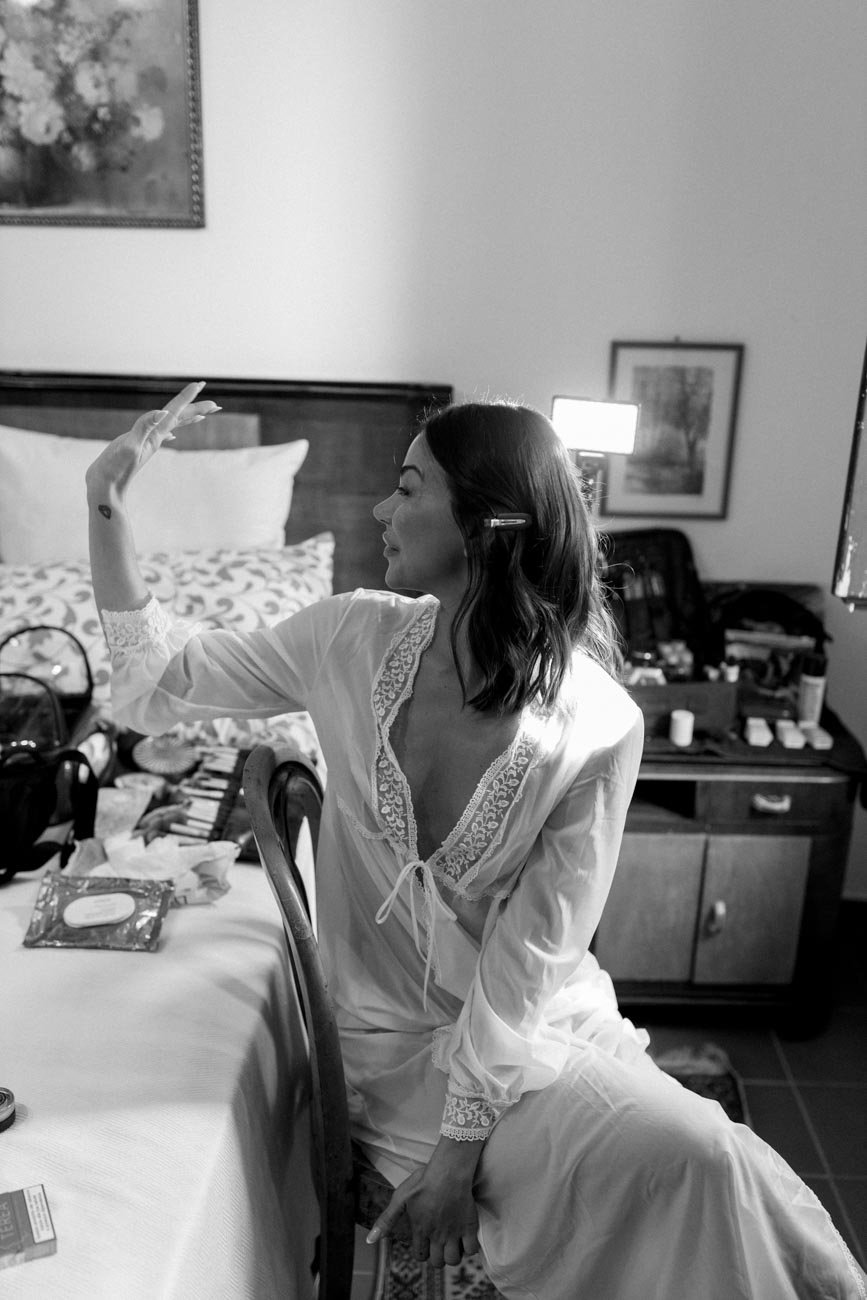 Una donna seduta su una sedia, indossa un abito bianco in pizzo, in un ambiente domestico. Sta facendo un gesto con la mano destra e sembra sorridere.