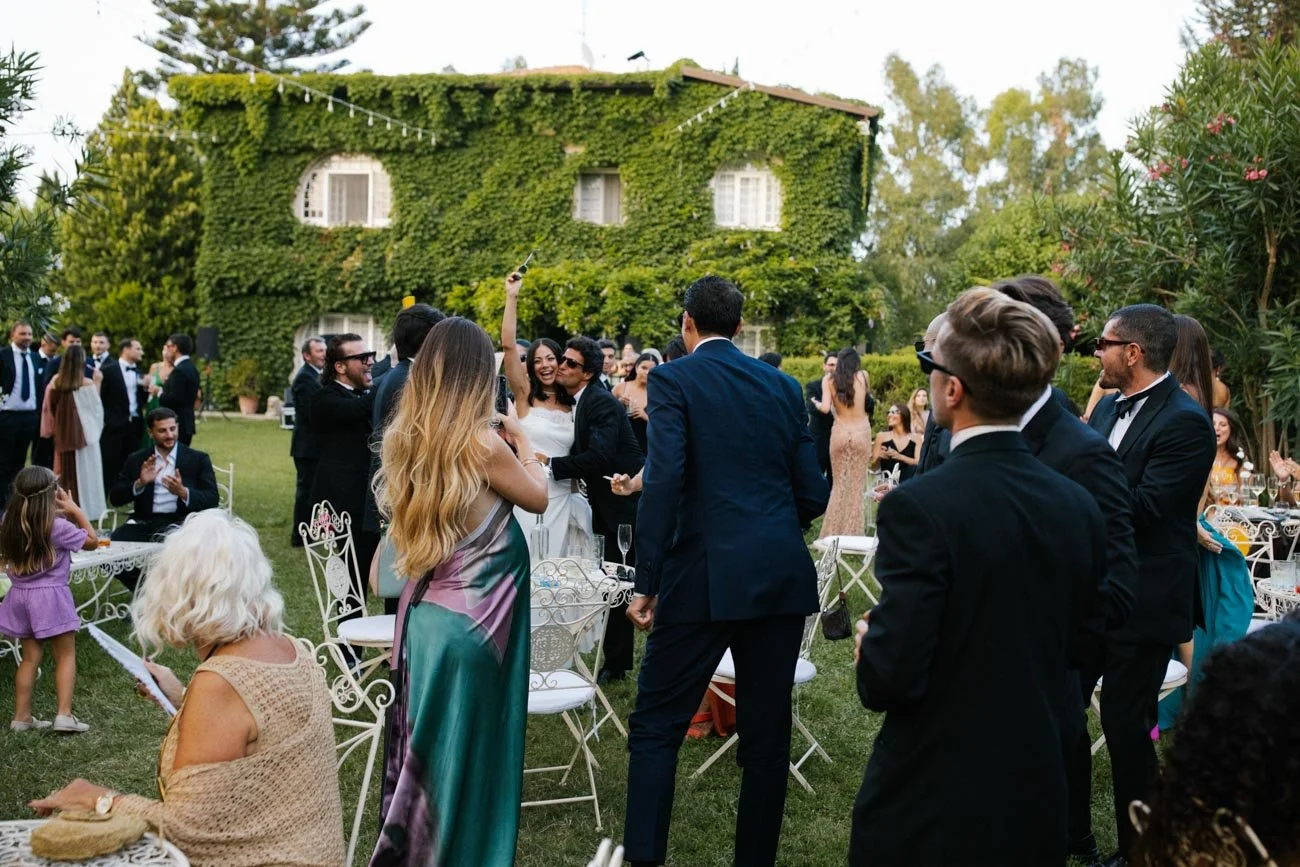 Celebrazione di matrimonio con persone vestite elegantemente, in giardino con edificio ricoperto di edera sullo sfondo.