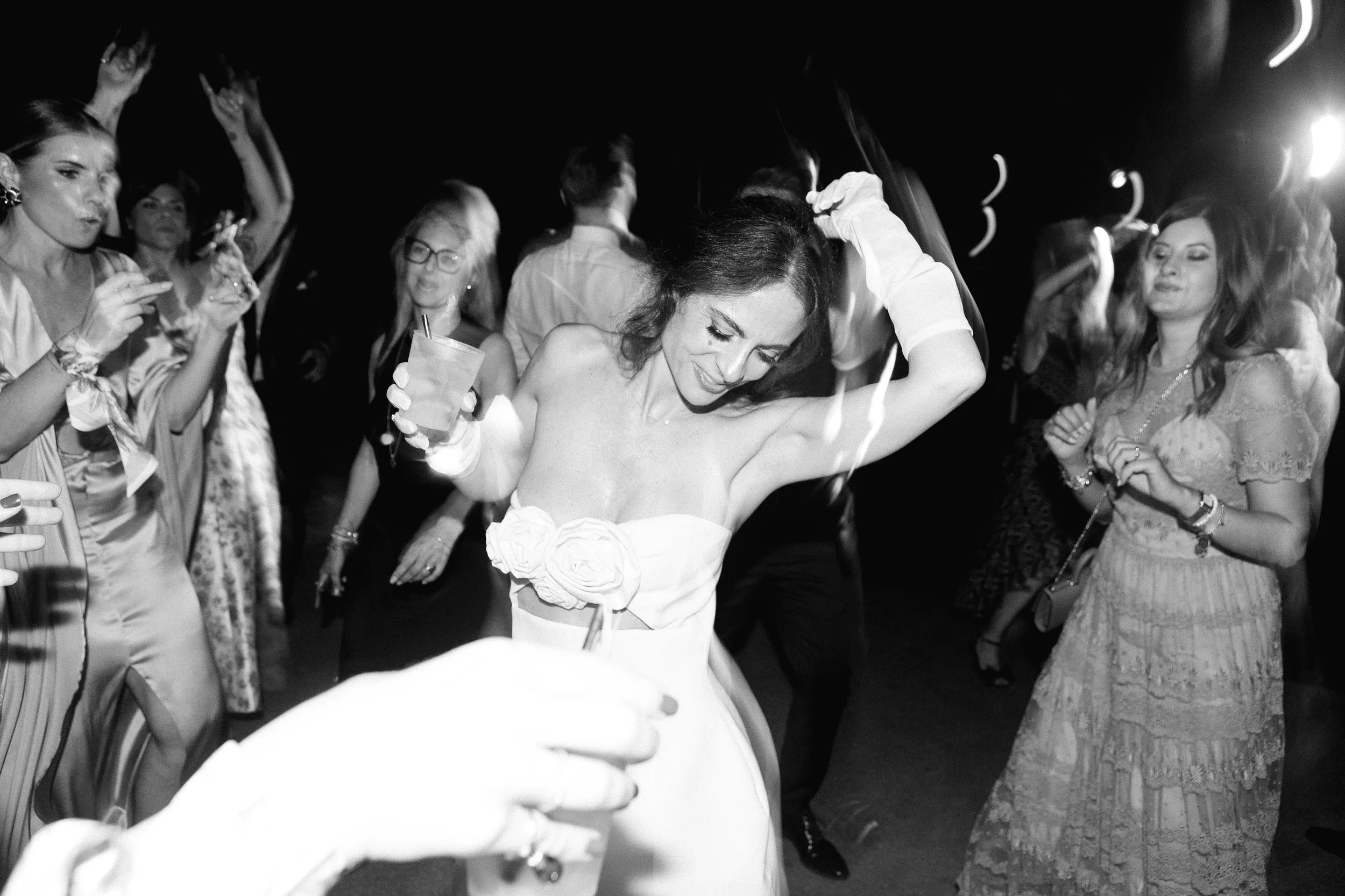 Una donna in abito bianco danza con un bicchiere in mano tra altre persone che festeggiano, in un ambiente party o cerimonia, in bianco e nero.