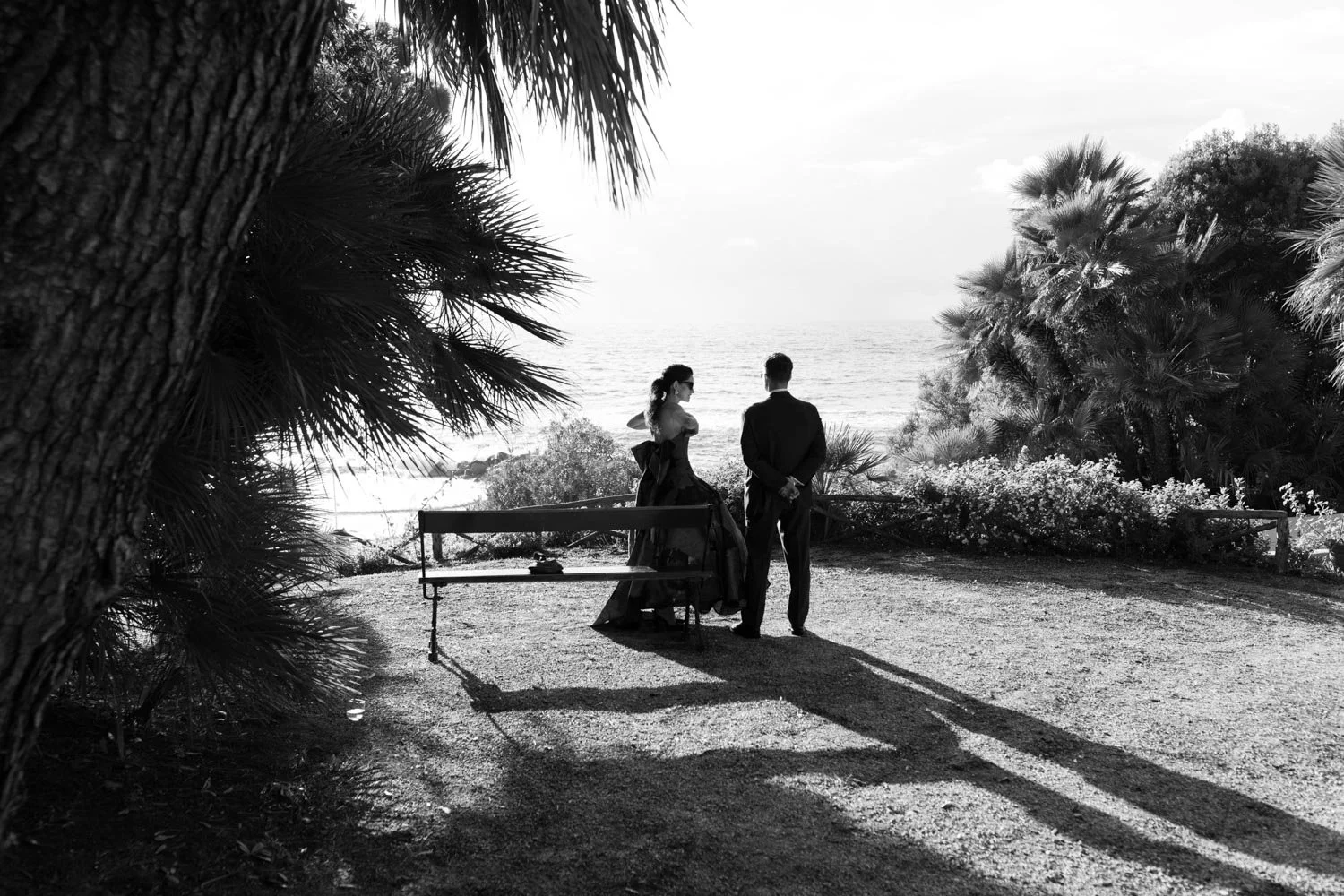 Due persone in abiti eleganti si incontrano su una spiaggia circondata da vegetazione, con il mare sullo sfondo, in una scena in bianco e nero.