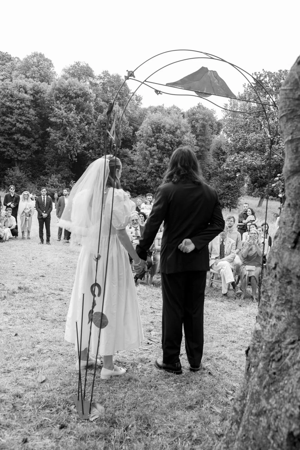 Una coppia si sposa in un matrimonio all'aperto, con la sposa in abito bianco e il volto coperto da un velo, e lo sposo in abito scuro, circondati da amici e parenti seduti e in piedi, con un paesaggio naturale sullo sfondo.