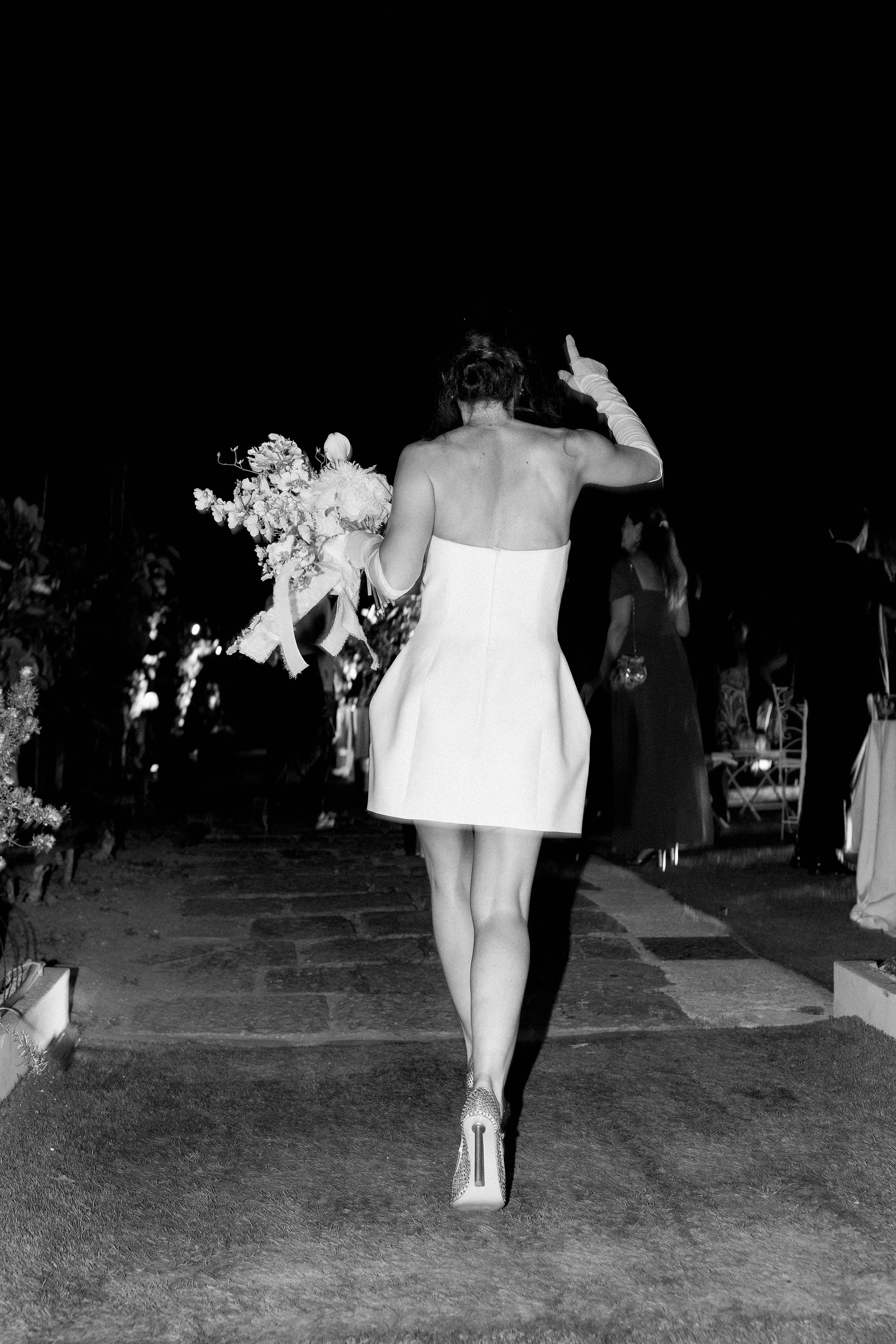 Donna in abito bianco che tiene un bouquet di fiori, vista di spalle, durante un evento serale.