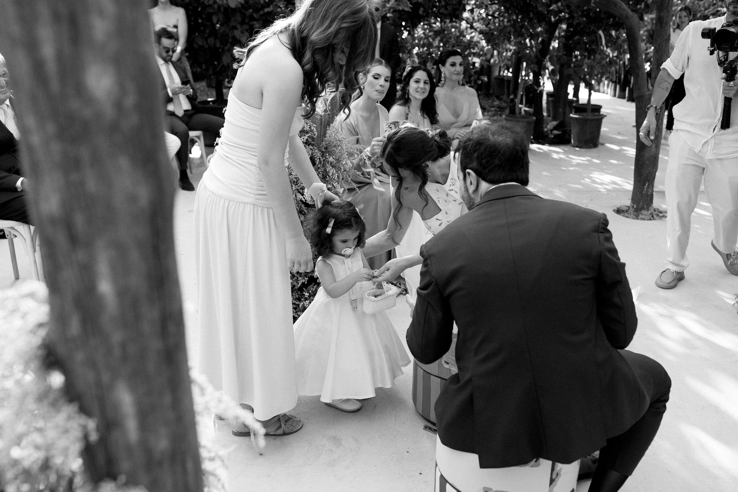 Coppia che si scambia gli anelli durante un matrimonio all'aperto, con persone sedute e alcune che osservano, in un ambiente naturale.