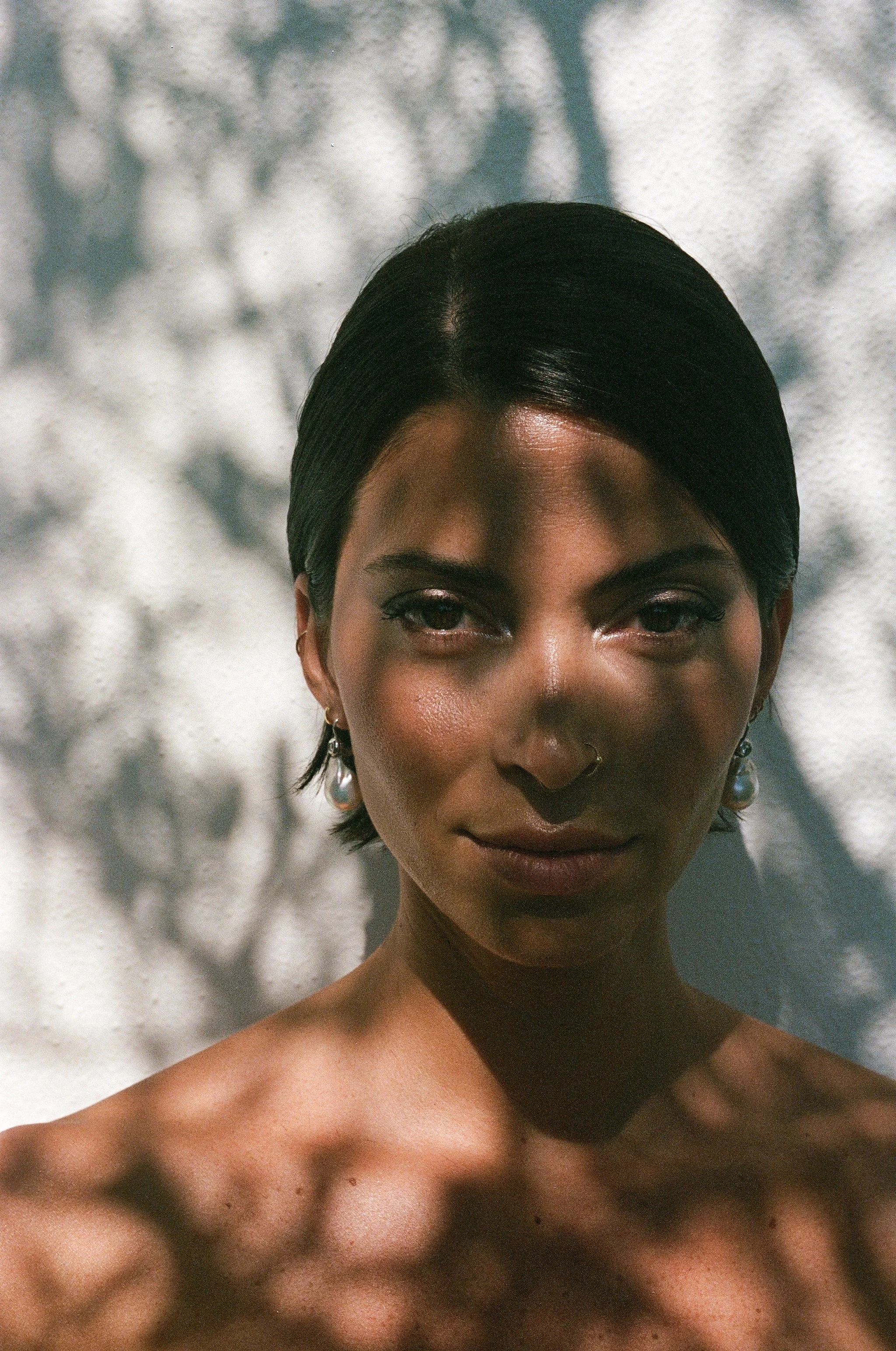 Ragazza con pelle scura e capelli neri lisci, con orecchini pendenti e un naso piercing, con un'illuminazione naturale che crea ombre di foglie sul suo volto e sulle spalle.