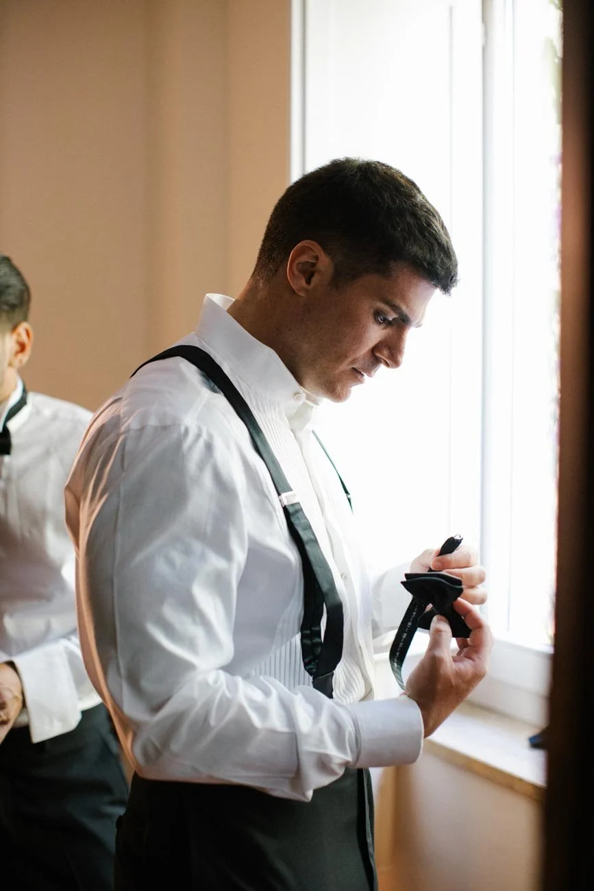 Uomo con camicia bianca e bretelle, che si sta preparando di fronte a una finestra, probabilmente un matrimonio o un evento formale.