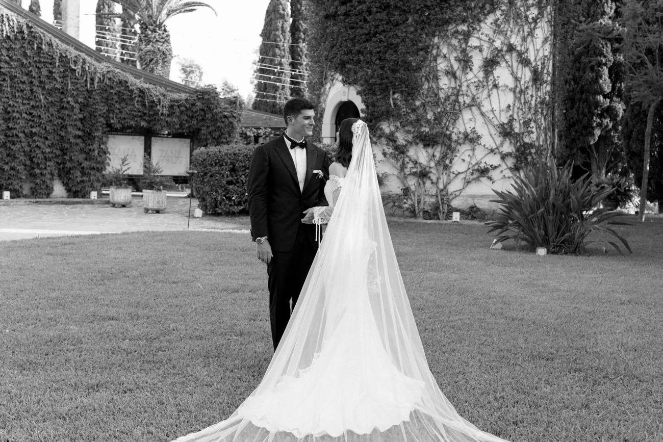 Una coppia di sposi si tiene per mano in un giardino, l'uomo indossa uno smoking e la donna un abito da sposa con velo.