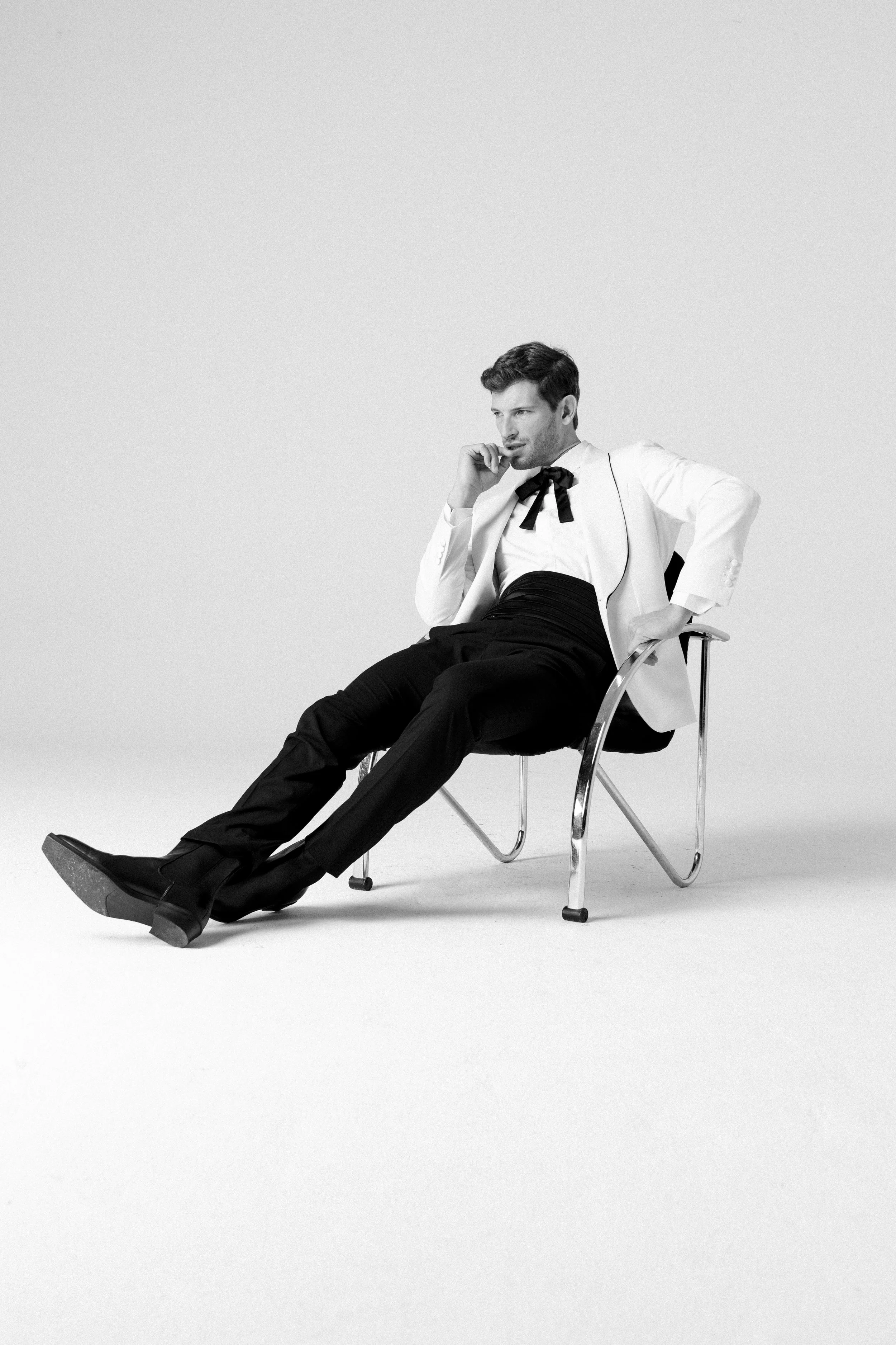 Uomo seduto su una sedia moderna, indossa abiti eleganti con giacca bianca, pantaloni neri e cravatta nera, in un'immagine in bianco e nero, pensieroso in un ambiente minimalista.