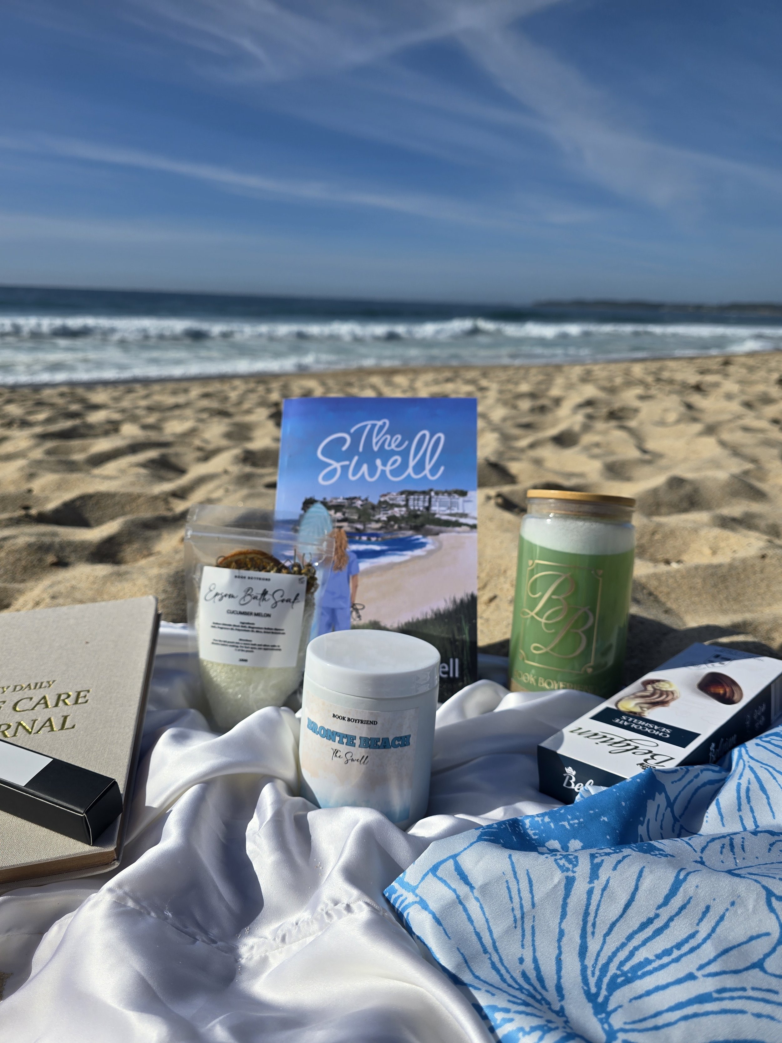 A collection of beach-themed items on sand, including a book titled 'The Swell', a jar labeled 'BLOUNTE BEACH', a container with a green label, a box of chocolates, a wrapped cloth with blue print, a white satin fabric, and a small box, with ocean waves and a clear sky in the background.