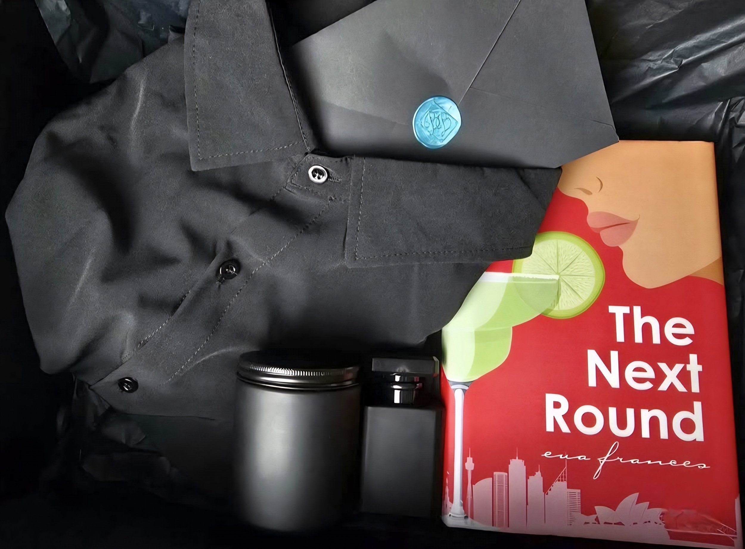 A black short-sleeve button-up shirt, a black envelope with a wax seal, a silver tumbler, a black flashlight, a colorful book titled 'The Next Round' by Eva Frances, and a black plastic bag inside a box.