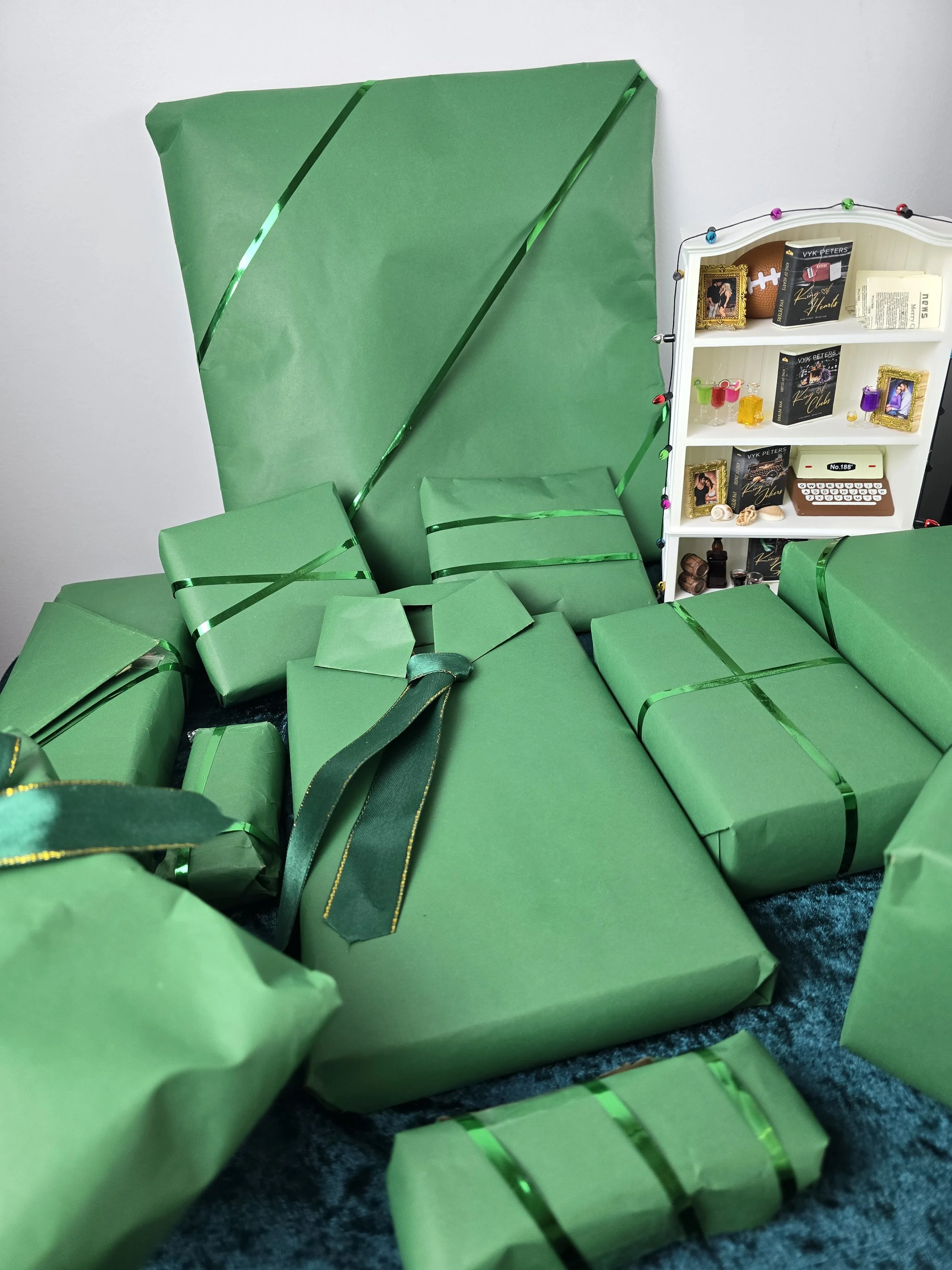 Multiple green wrapped Christmas presents of various sizes on a blue carpet, with a large gift leaning against the wall in the background, and a white shelf decorated with small framed photos, books, and collectibles to the right.