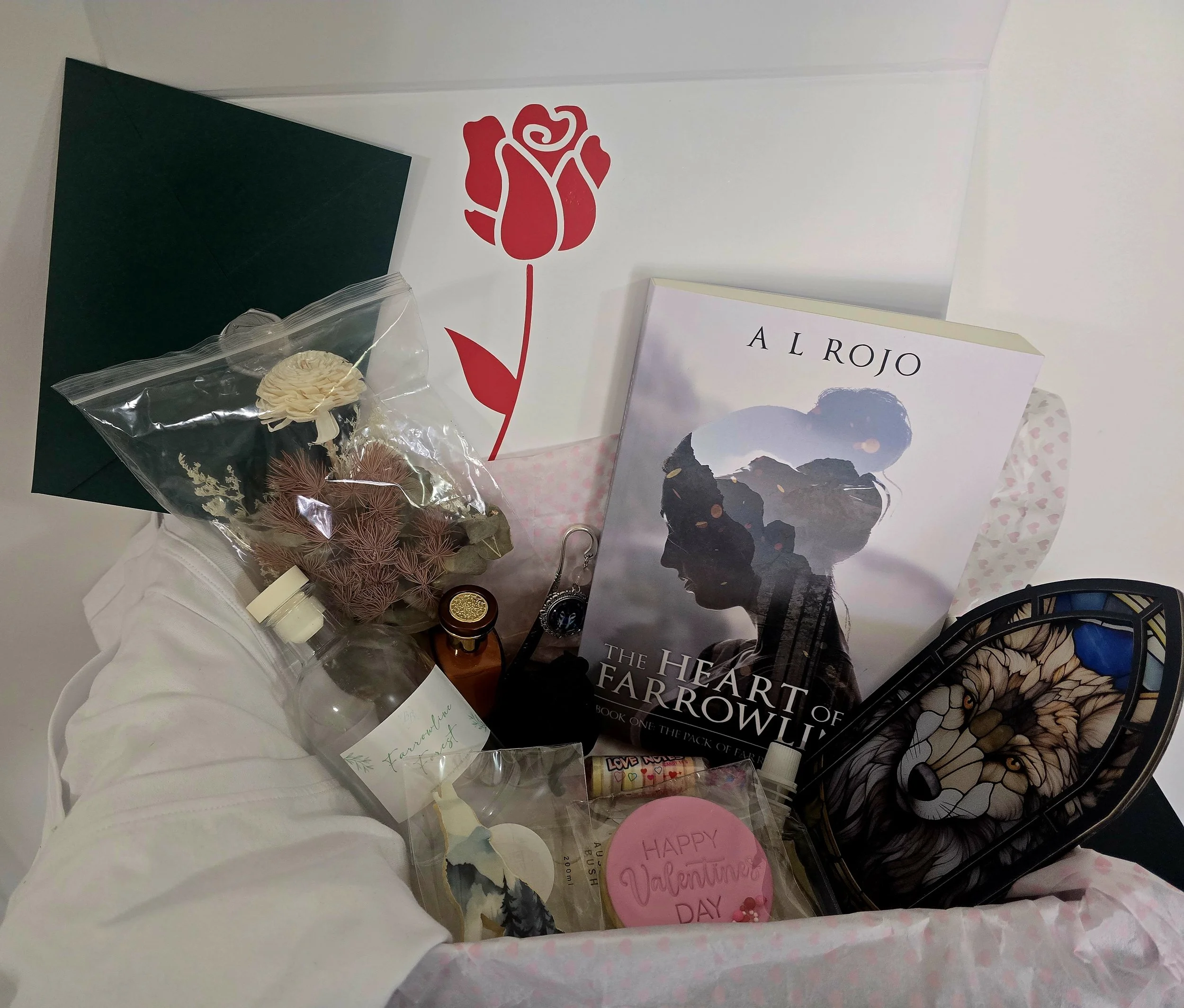 A gift basket containing a book titled 'The Heart of Farrowly' by Al Rojo, a Valentine's Day card, a small glass bottle, dried flowers, a packaged heart-shaped item, and a sketch of a wolf's face on a decorative box.