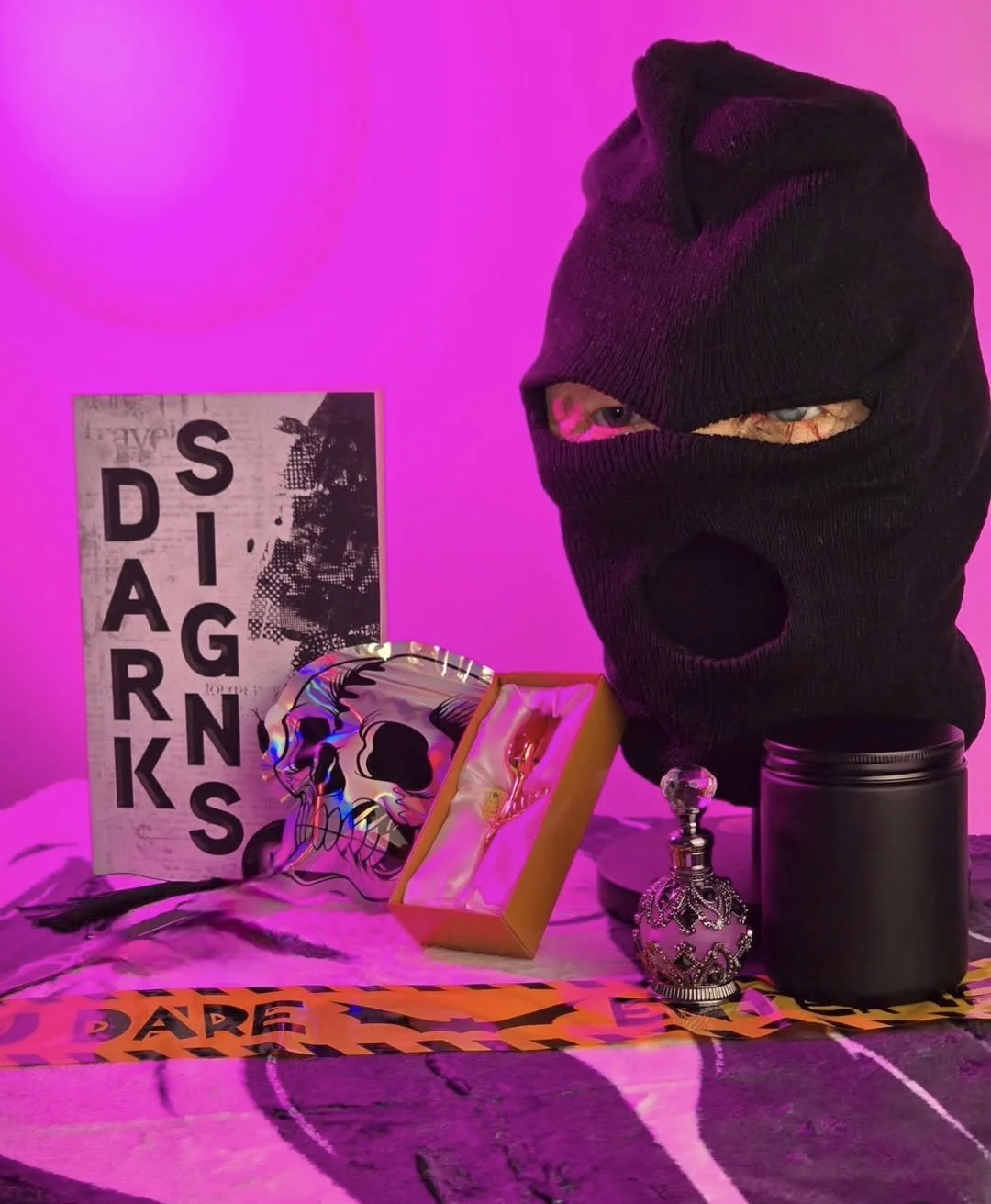 A table with Halloween themed items, including a black ski mask with eye and mouth holes revealing eyes and parts of a face, a black candle, a decorative skull-shaped bottle, a viper in a box, and a poster with the words "DARK SIDE GRAPHICS" on a pink-lit background.