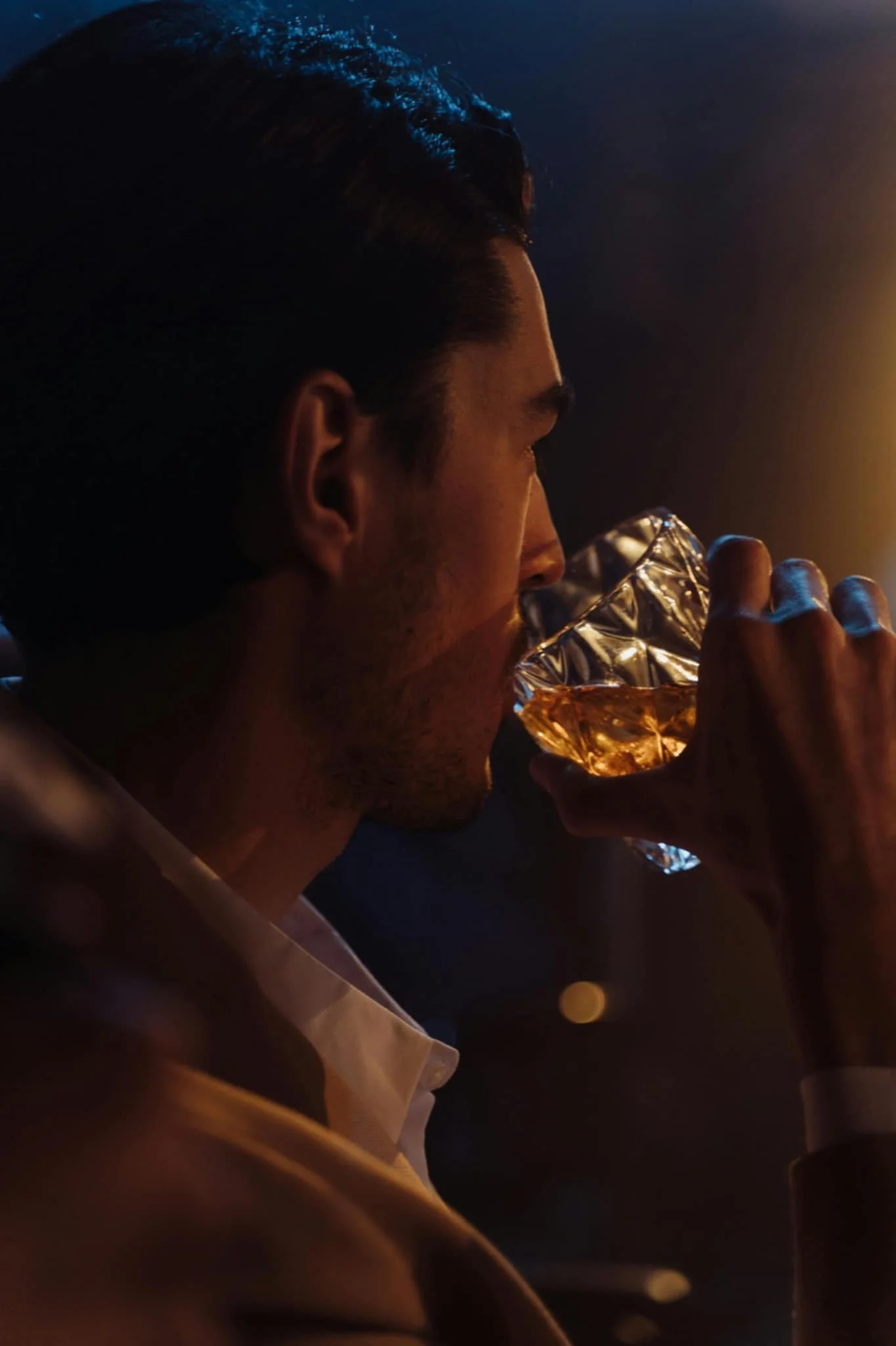 A man with dark hair and a light shirt is drinking whiskey from a textured glass in a dimly lit setting.