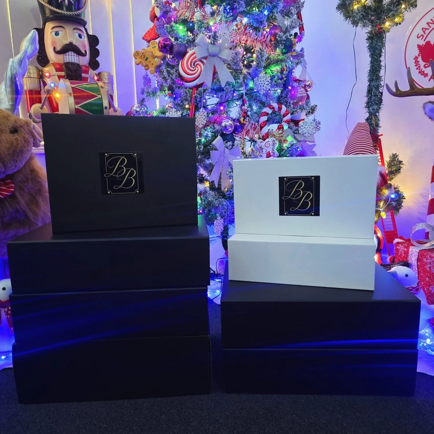 Four large shopping bags, two black and two white, with a logo 'BB' on them, displayed in front of a decorated Christmas tree with colorful lights, ornaments, and candy canes. Christmas decorations and plush toys are also visible in the background.