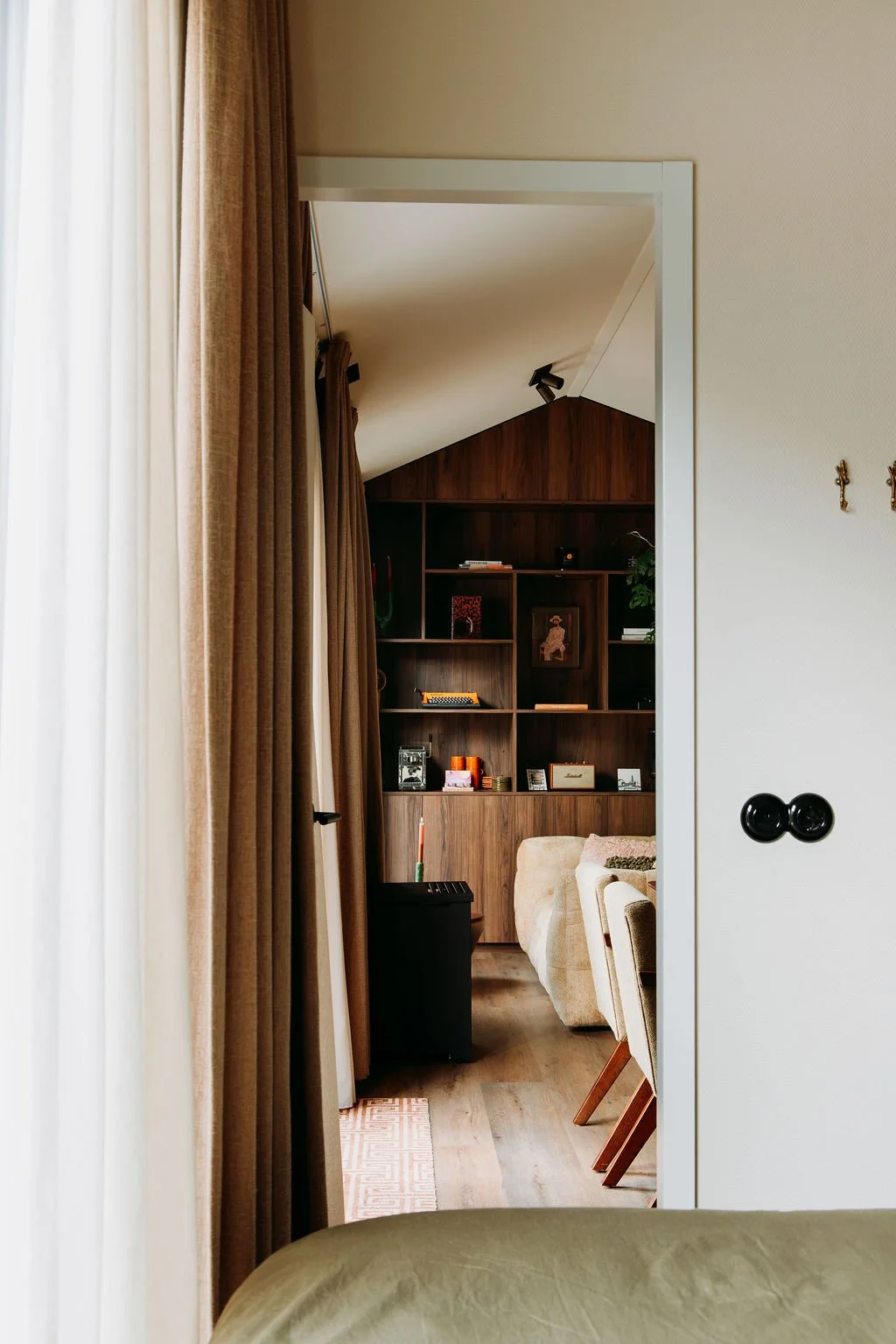 Een interieurzicht door een deuropening met gordijnen links, een beige bed in de voorgrond, een witte muur met zwarte schakelaars van De Oude Schakelaar, en een woonkamer met een houten kast en boekenplanken, enkele stoelen van Loods 5.