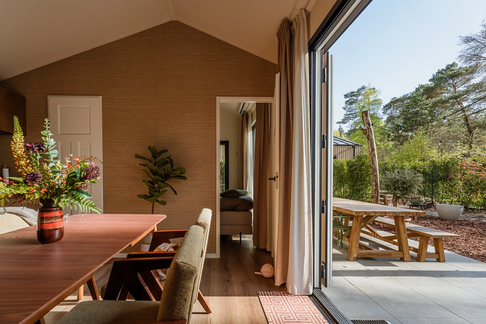 Interieur van een huis met een eettafel met een boeket bloemen en een uitzicht op de tuin via open deuren.