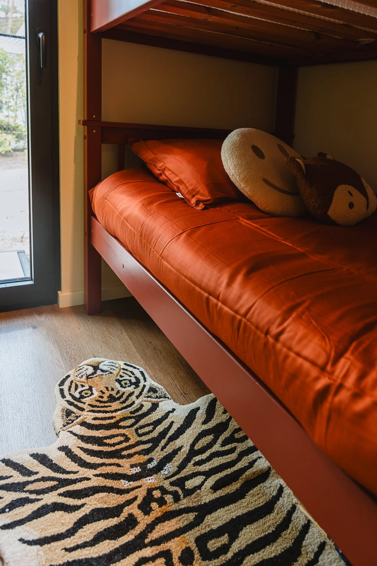 Een bonten tapijt met een tijgerprint ligt op de vloer onder een bed met oranje beddengoed en twee kussens, een met een glimlachgezicht en de ander met een koehoofd, naast een deur met een glasvenster.