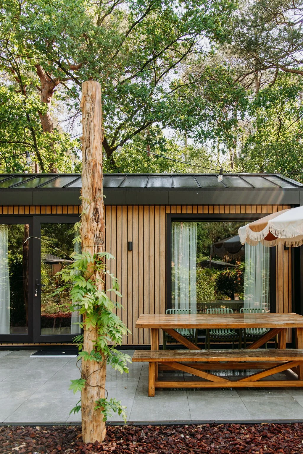 Tuin van een tiny house, een houten tafel, een parasol, een boom, groene bladeren, en een betegeld terras.