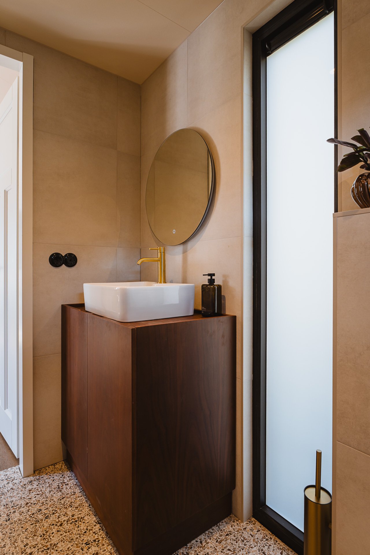 Moderne badkamer met ronde spiegel, witte wastafel op houten kast, goudkleurige kraan, zwart zeepdispensertje, en een frosted glas deur.