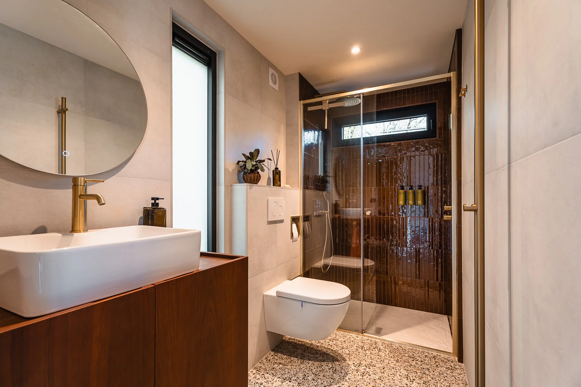 Moderne badkamer met witte wastafel, spiegel, roestvrijstalen kraan, wandcloset, inloopdouche met glazen schuifdeur en donkere tegels.