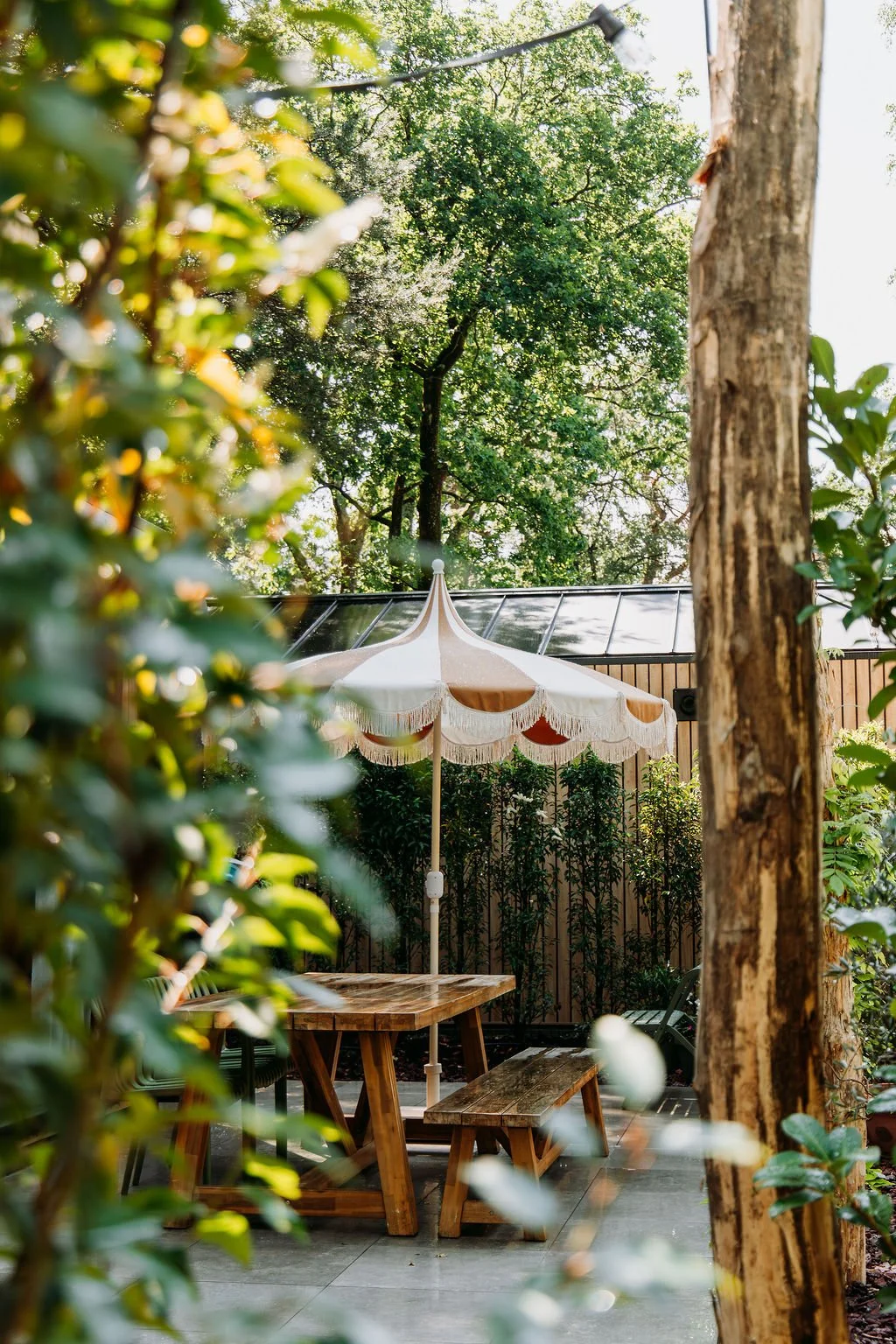 Een tuin met een houten tafel, bank en parasol, omgeven door groene bomen en struiken.