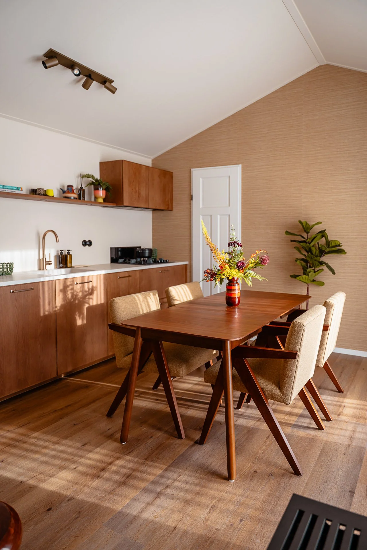 Een eetkamertafel met vier beige stoffe kuipstoelen en een vaas met bloemen in het midden. Achter de tafel bevindt zich een keuken met houten kasten en een wit werkblad, boven het werkblad een open plank met decoratieve items en planten. De muur aan 