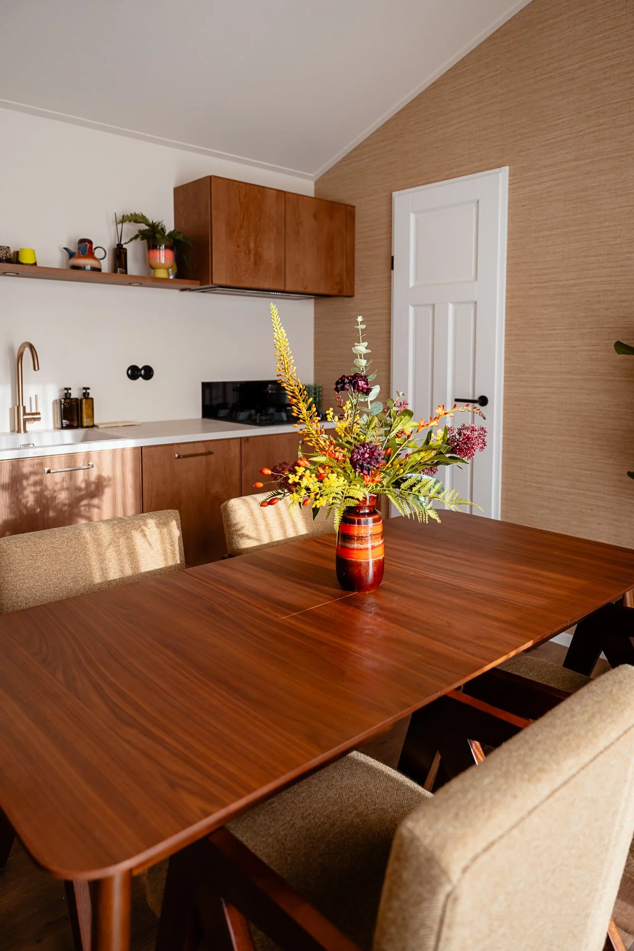 Een middelpunt met een bloemenvaas op een houten eettafel, op de achtergrond een keuken met houten kasten en een witte deur.