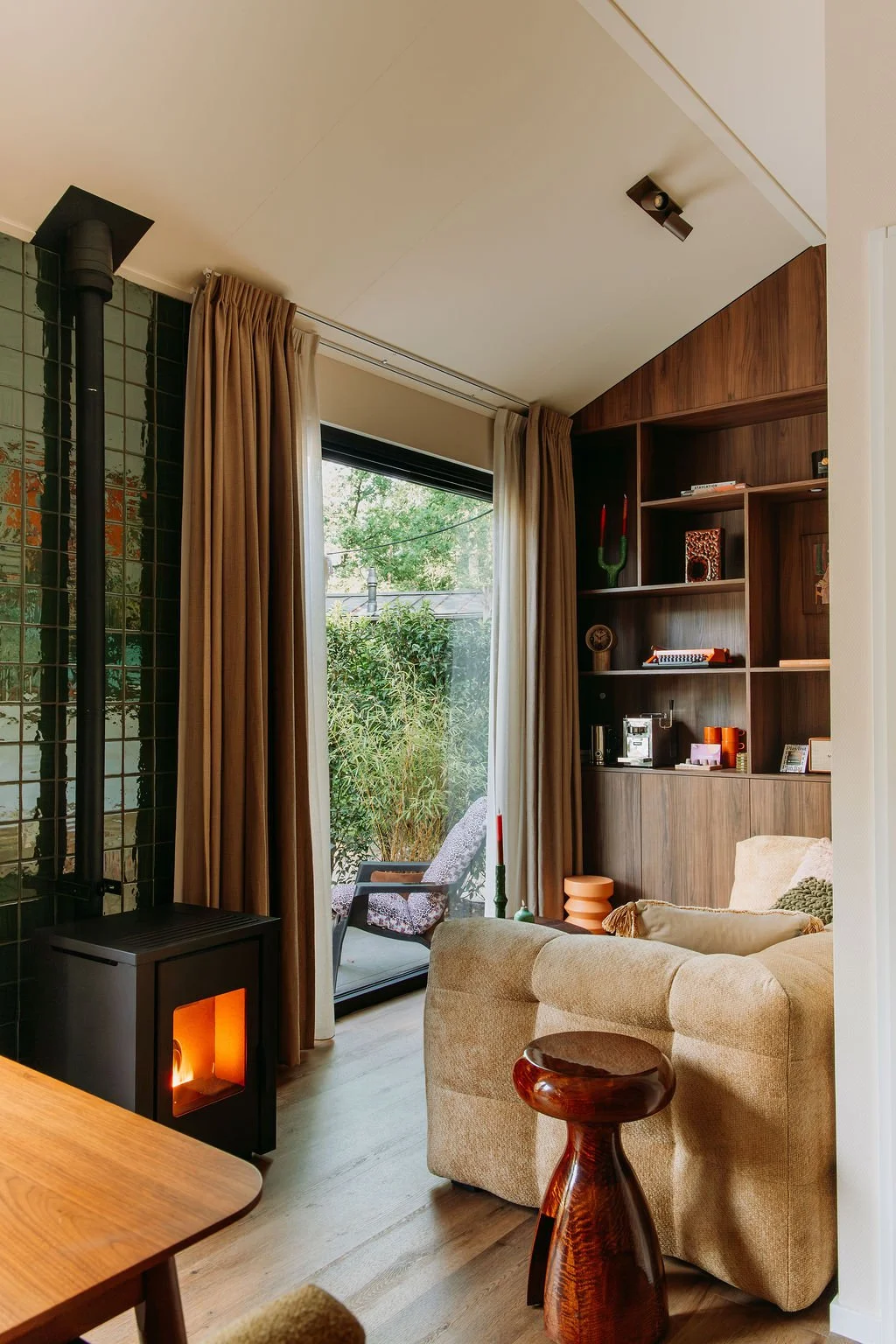 Een gezellige woonkamer met een open haard, grote ramen met beige gordijnen, een boekenkast van hout met decoratieve spullen, een LOODS 5 bank en een HKLiving houten kruk.