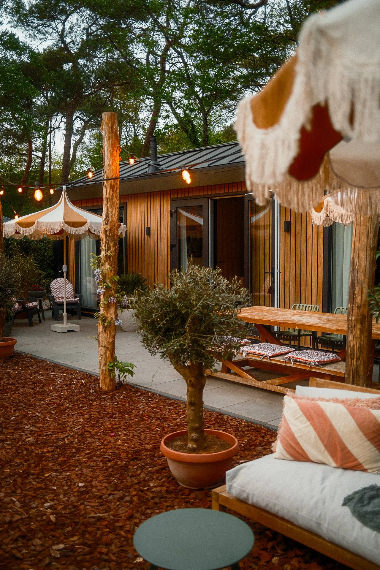 Een gezellige patio met houten meubelen, een parasol, planten, tuinlampjes en een houten huis met grote ramen op een avond.